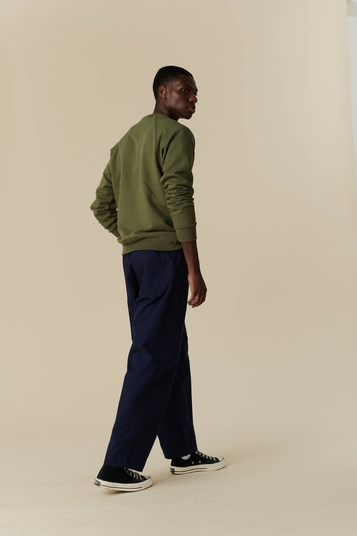 
            Back angle picture of a man wearing olive sweatshirt with navy tapered fit cameraman pant with black 70s Converse.