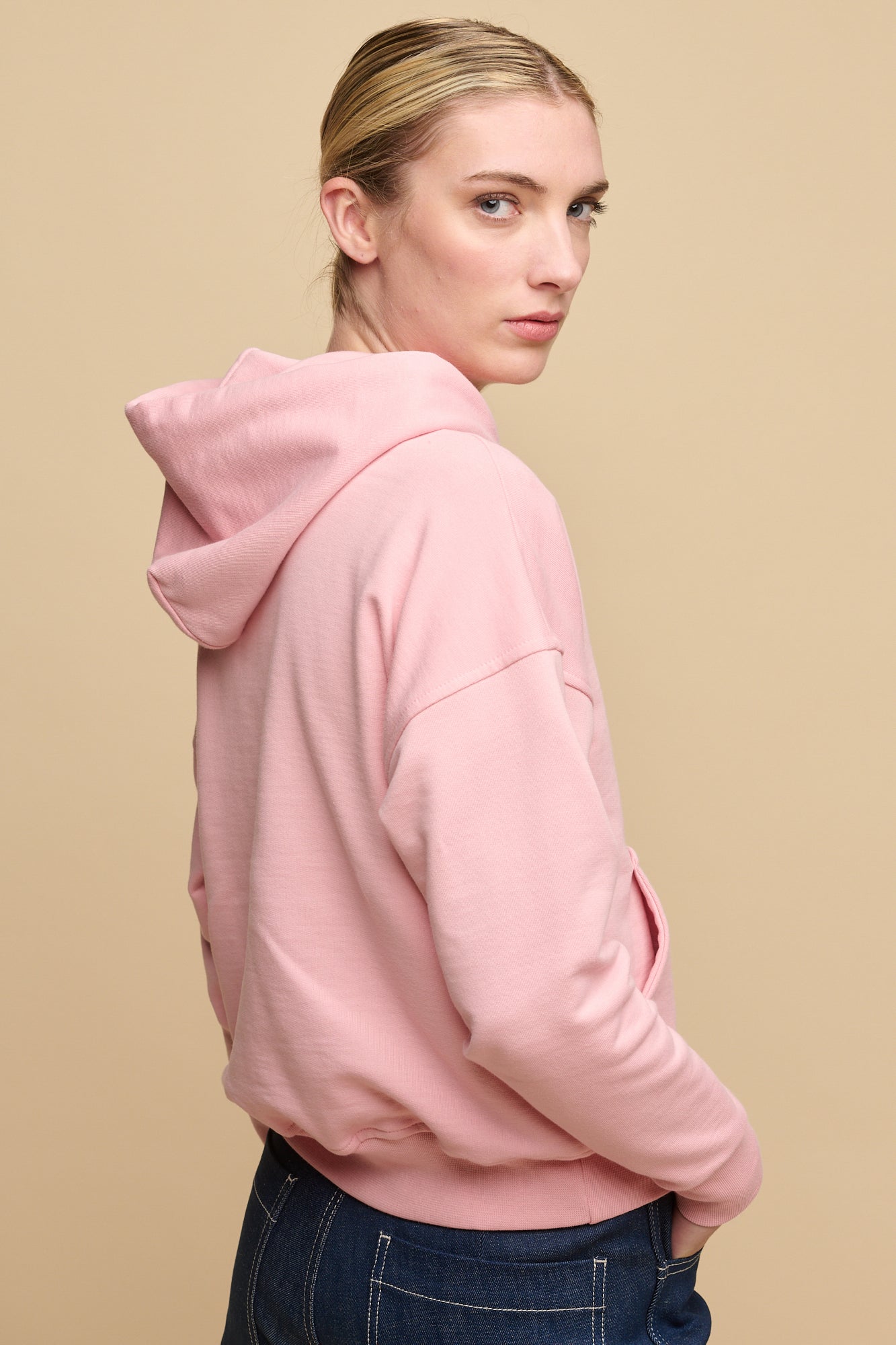 Thigh up image of the back of female wearing pale pink hooded sweatshirt paired with work jeans in blue