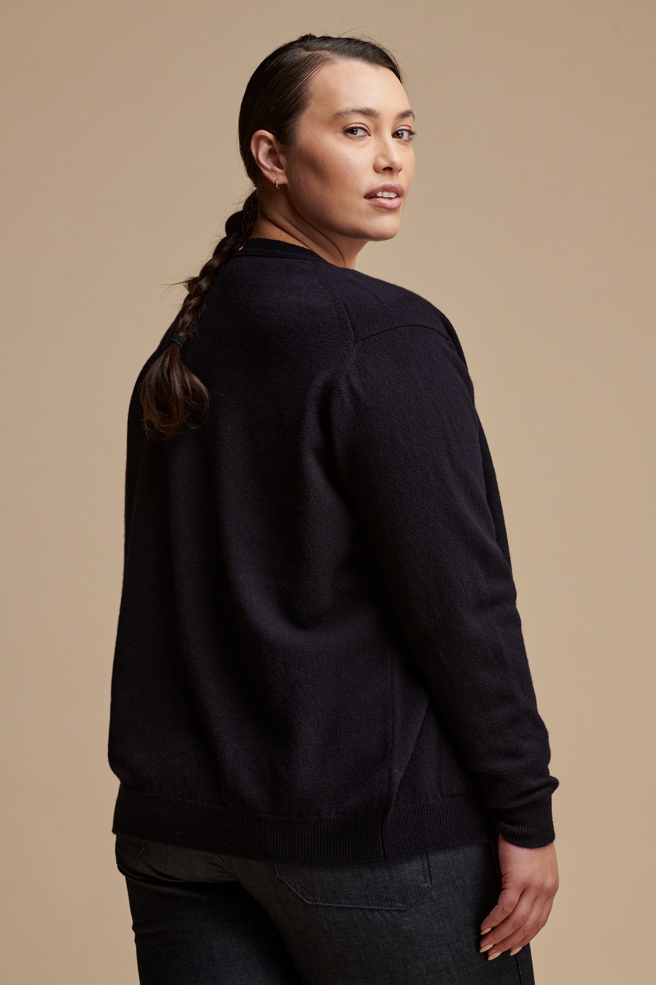 Thigh up image of the back of female with her hair tied in a low plait wearing lambswool v neck cardigan in navy paired with work trousers in indigo.