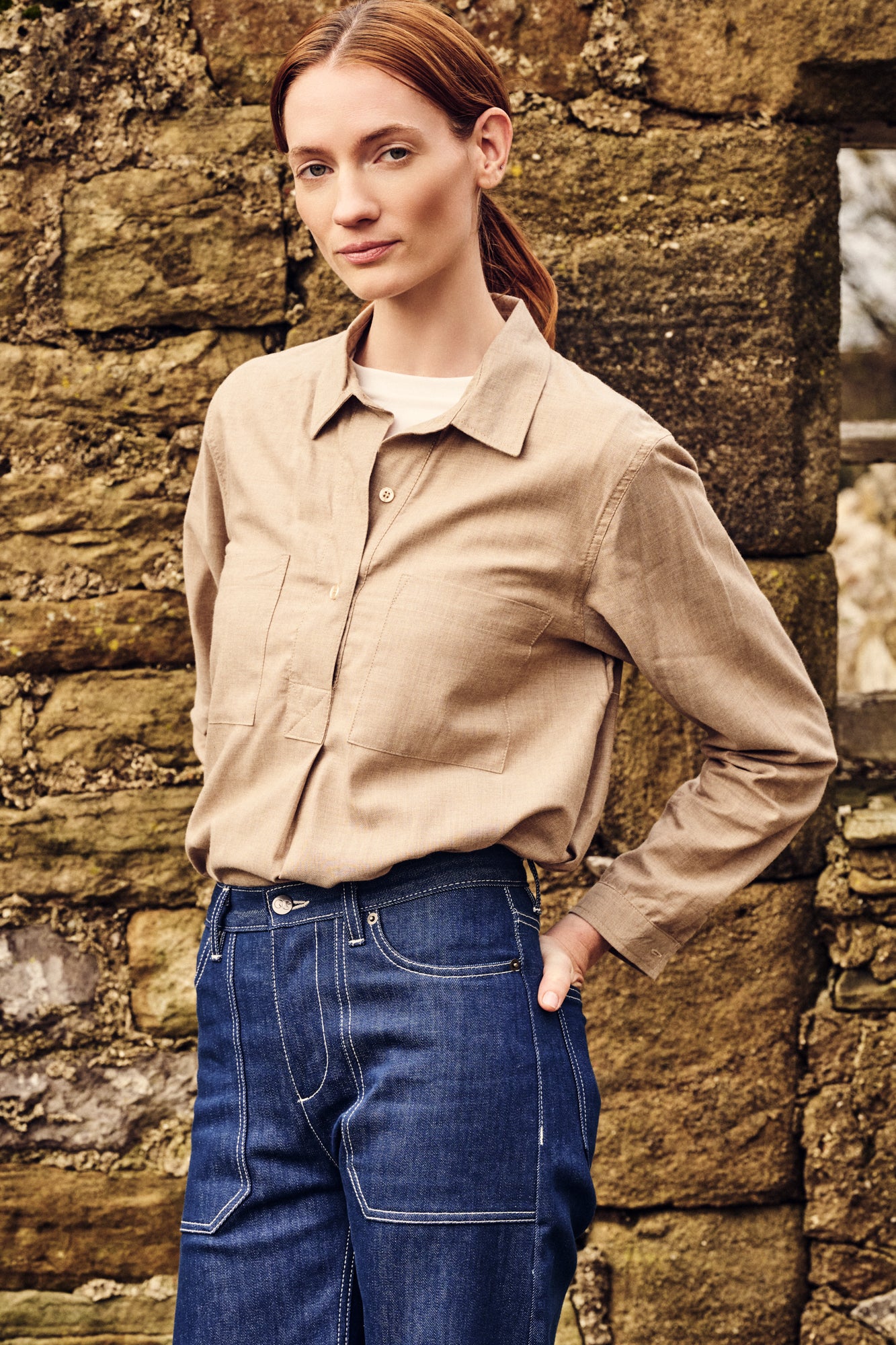 Woman wearing a beige shirt and blue jeans standing against a stone wall.