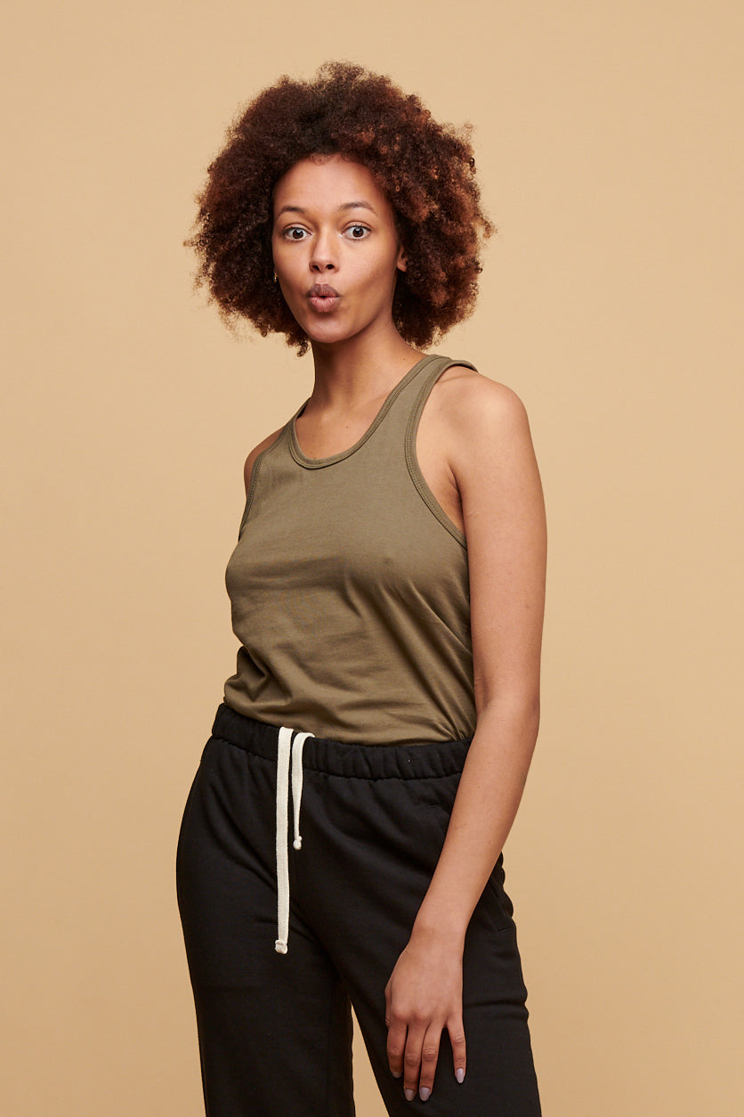 Female from the thigh up wearing racer back vest in olive made form 100% cotton tucked into black sweatpants also made from 100% cotton