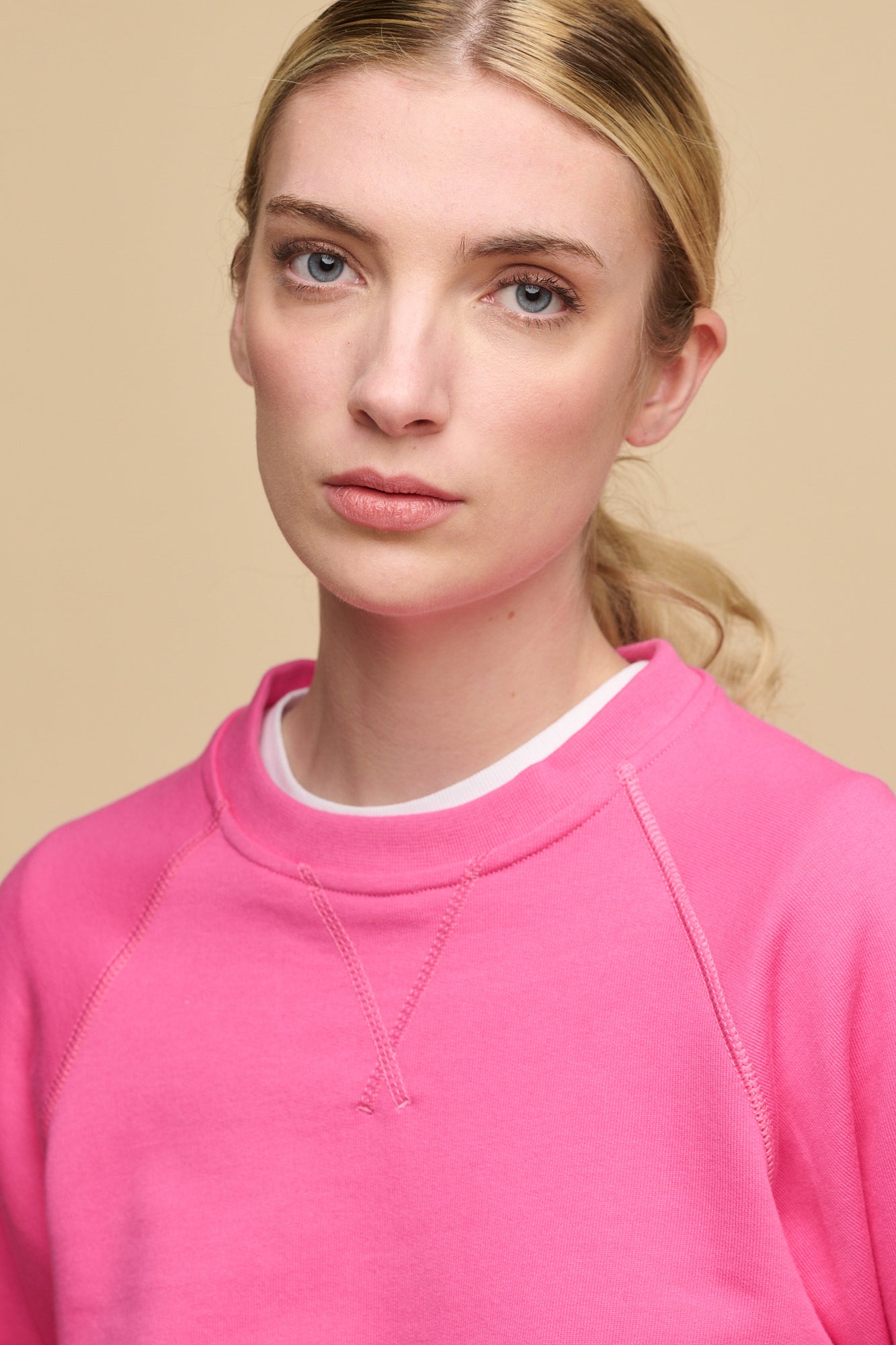 Head shot of bkonde female with blue eyes wearing raglan sweatshirt in fuchsia pink worn over crew neck t shirt in white