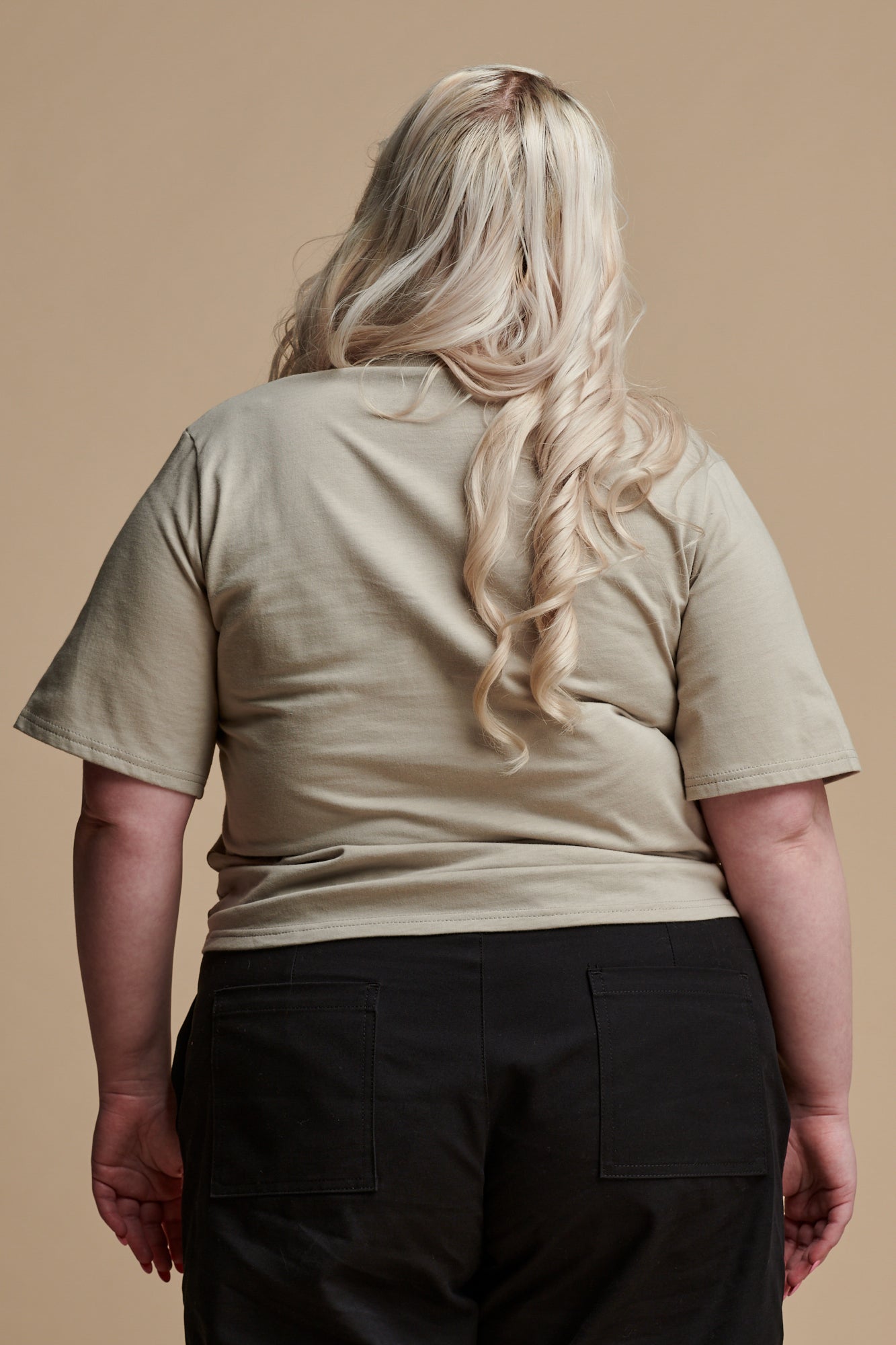 Thigh up of the back of female with long, curled blonde hair wearing short sleeve v neck t shirt in stone paired with camerawoman pants in black.