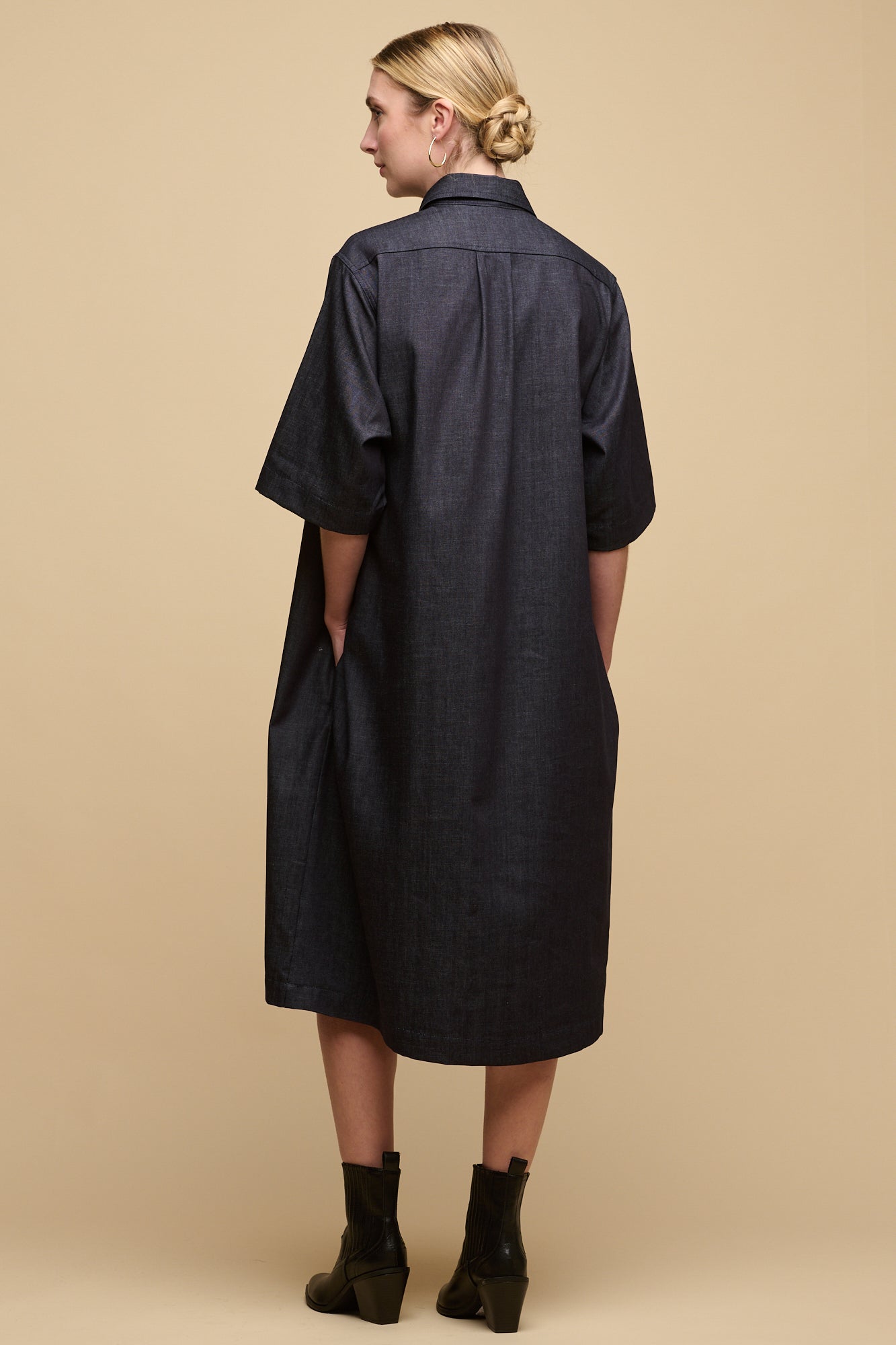 Back of female wearing untied straight denim dress in indigo with hands in pockets