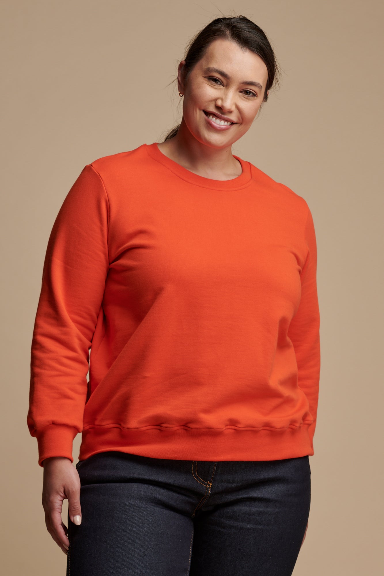 Thigh up of smiley female with dark hair tied back wearing sweatshirt in flame red paired with work jeans in indigo