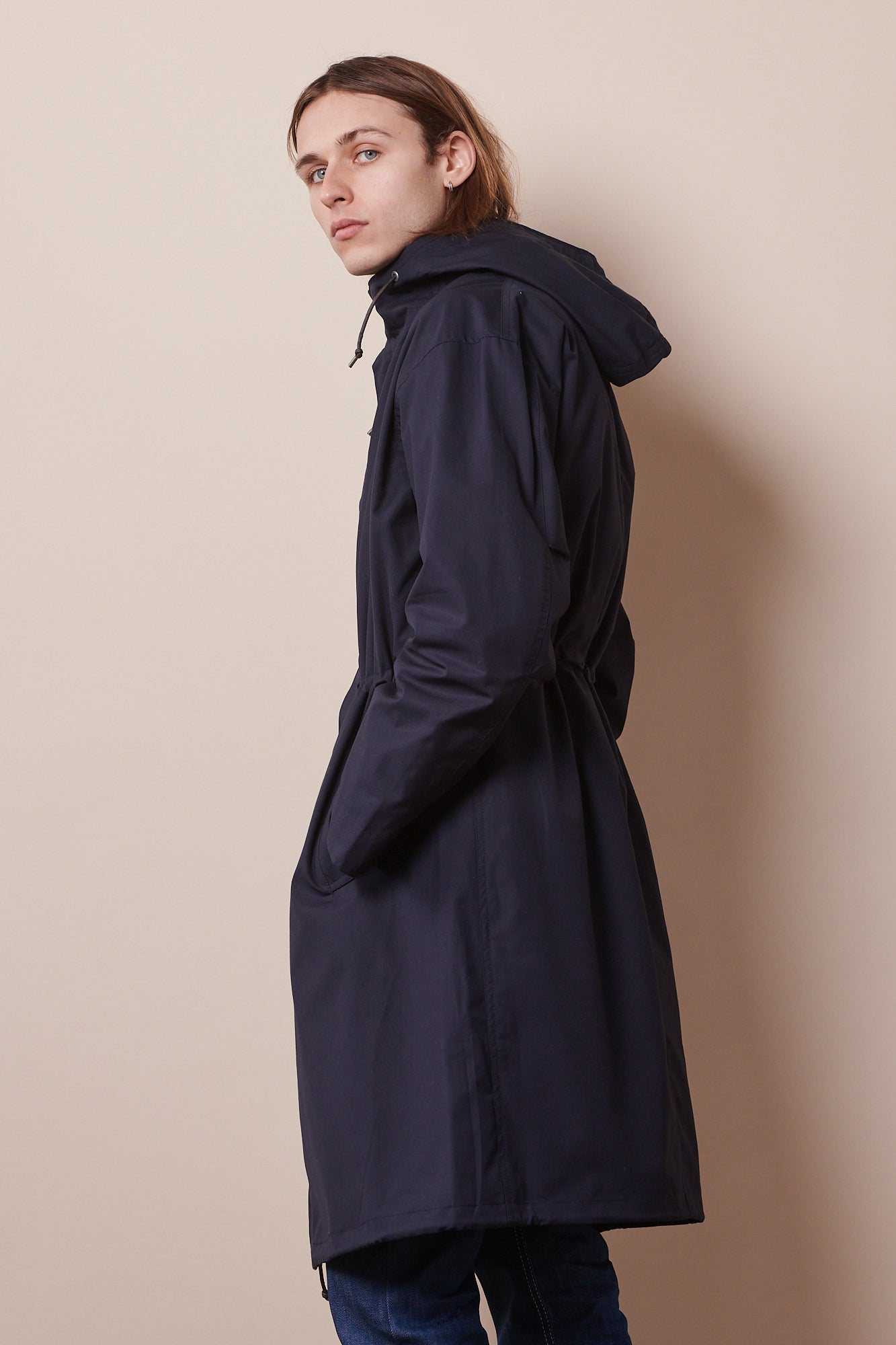 Back of male looking back towards camera wearing navy parka with hands in pockets. Adjustable waist cord tightened to pull waist in