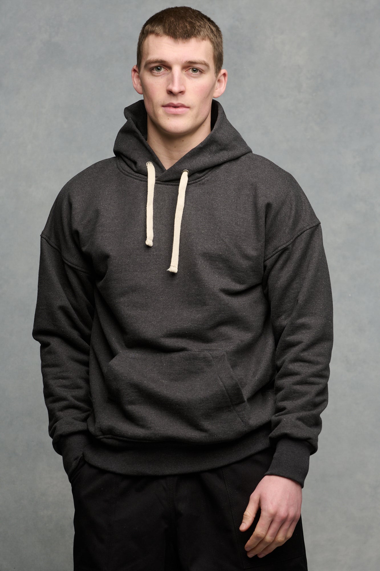 Thigh up front of brunet male wearing heritage hooded sweatshirt in charcoal paired with black cameraman pants