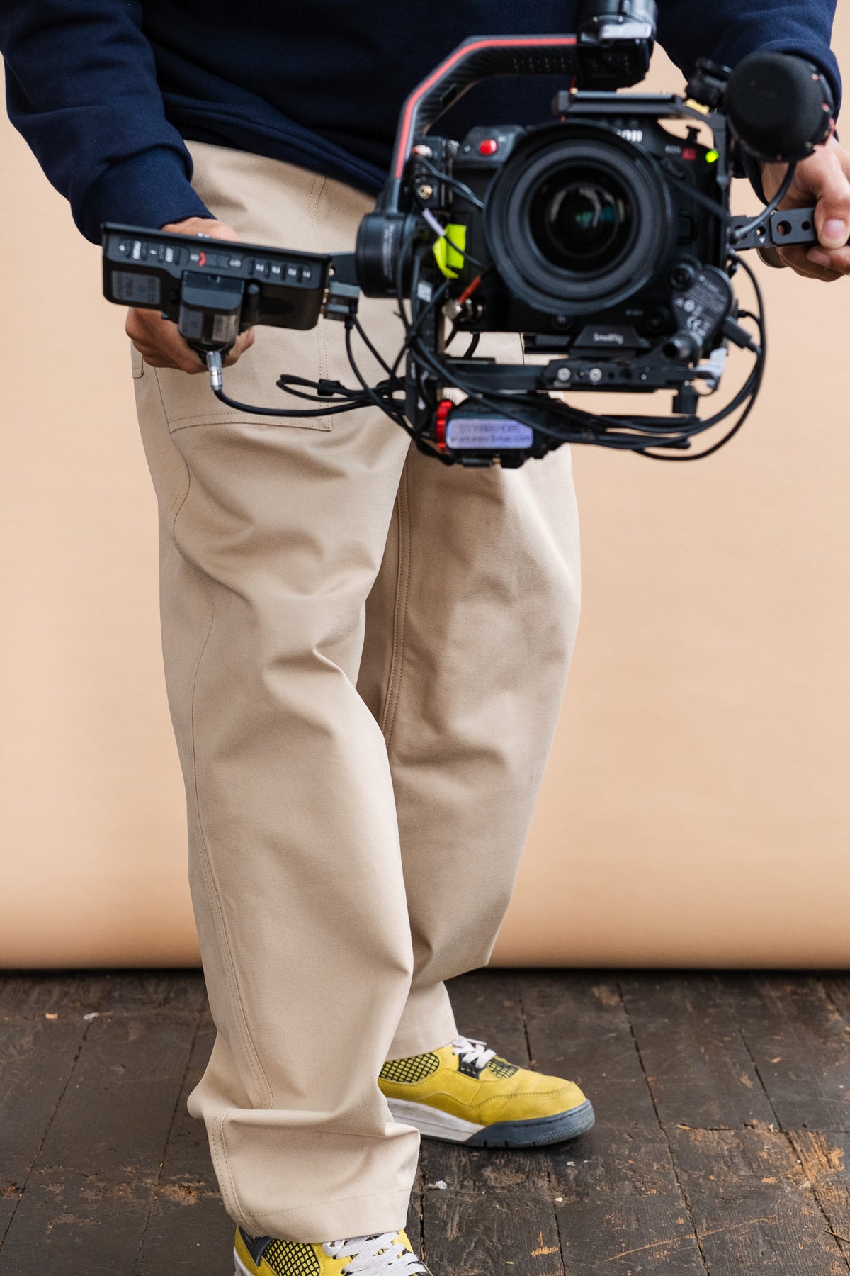 person holding a camera wearing a navy sweatshirt and the camerman pant in putty stood on wooded flooring in front of a beige backdrop