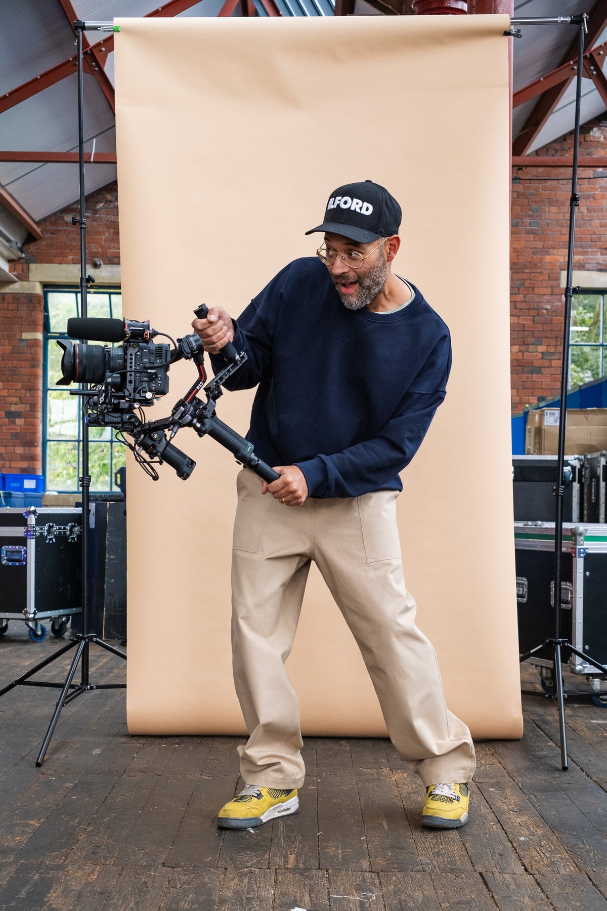 
            male wearing putty cameraman pant and a navy sweatshirt whilst holding a camera in front of a beige background