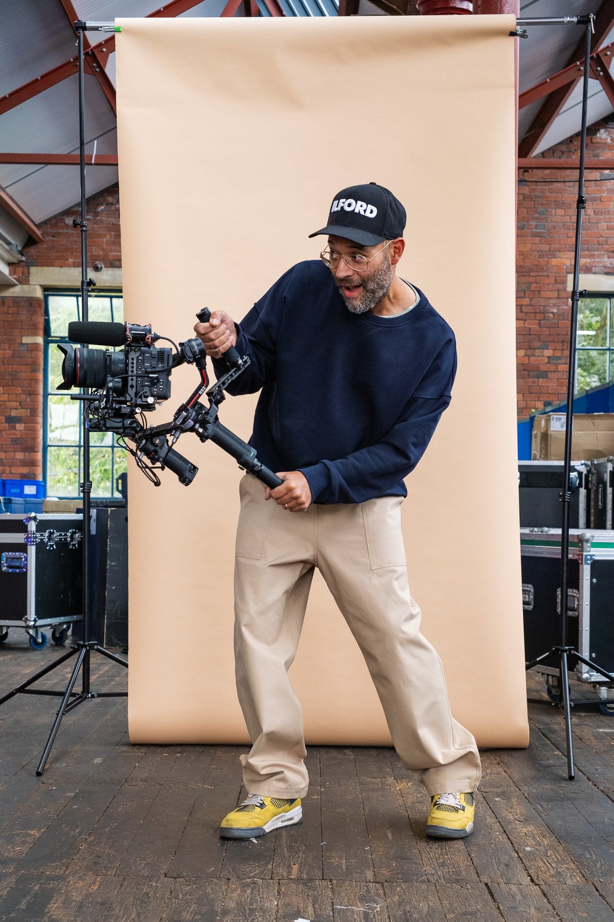 male wearing putty cameraman pant and a navy sweatshirt whilst holding a camera in front of a beige background