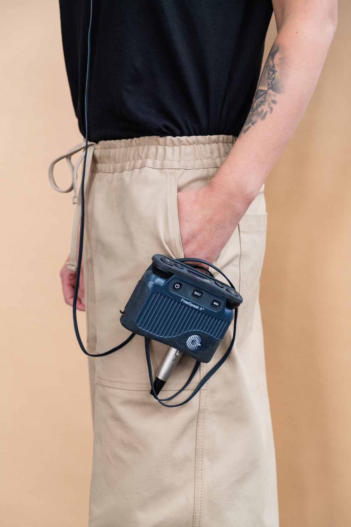 Close up of a person wearing a black t-shirt tucked into the cameraman pant in putty with their left hand in their pocket and a recorder attached to the pocket