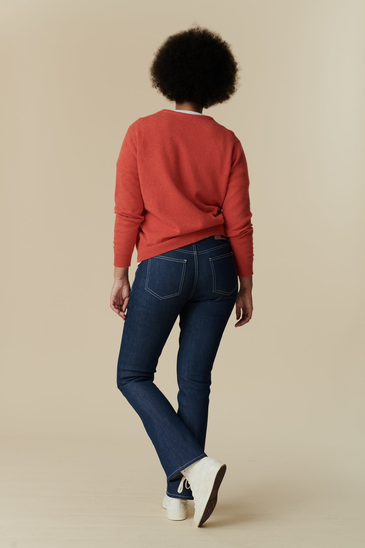 Full body back shot of black female model with afro wearing the women's bootcut mid rise jeans in blue paired with flame red, lambswool crew neck jumper