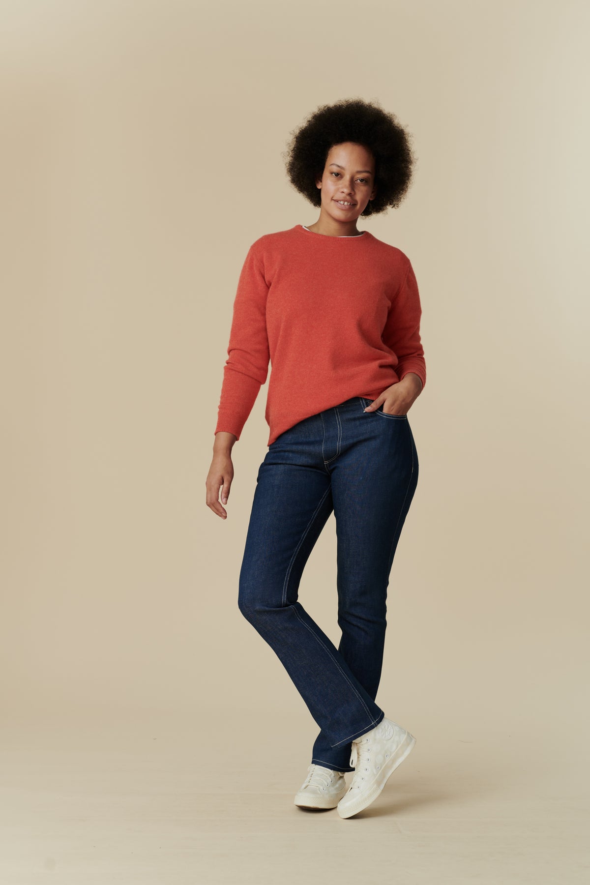 Full body front shot of black female model with afro wearing the women's bootcut mid rise jeans in blue paired with flame red, lambswool crew neck jumper