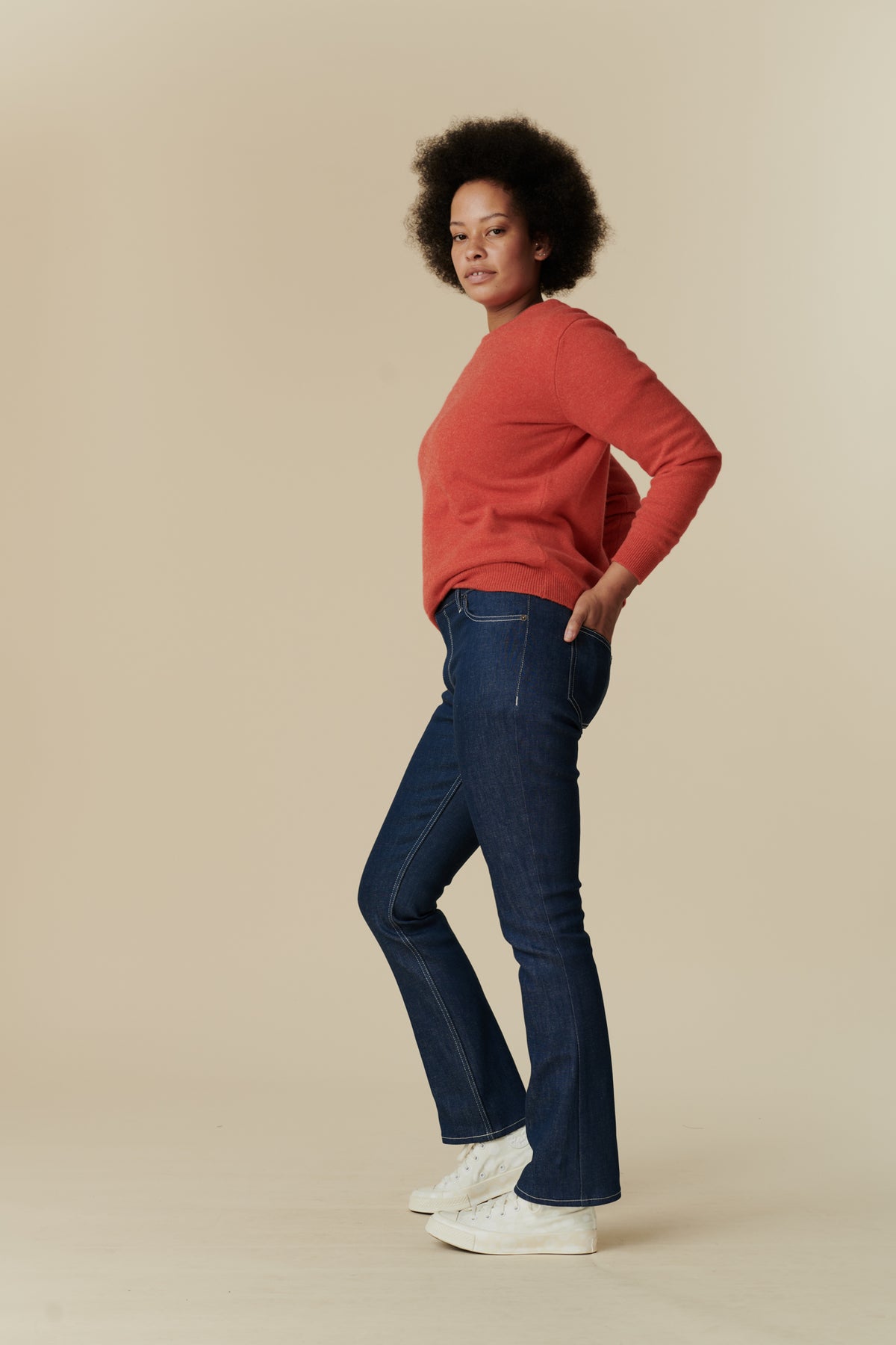 
            Full body side shot of black female model with afro stood with hands in back pockets of the women&#39;s bootcut mid rise jeans in blue paired with flame red, lambswool crew neck jumper