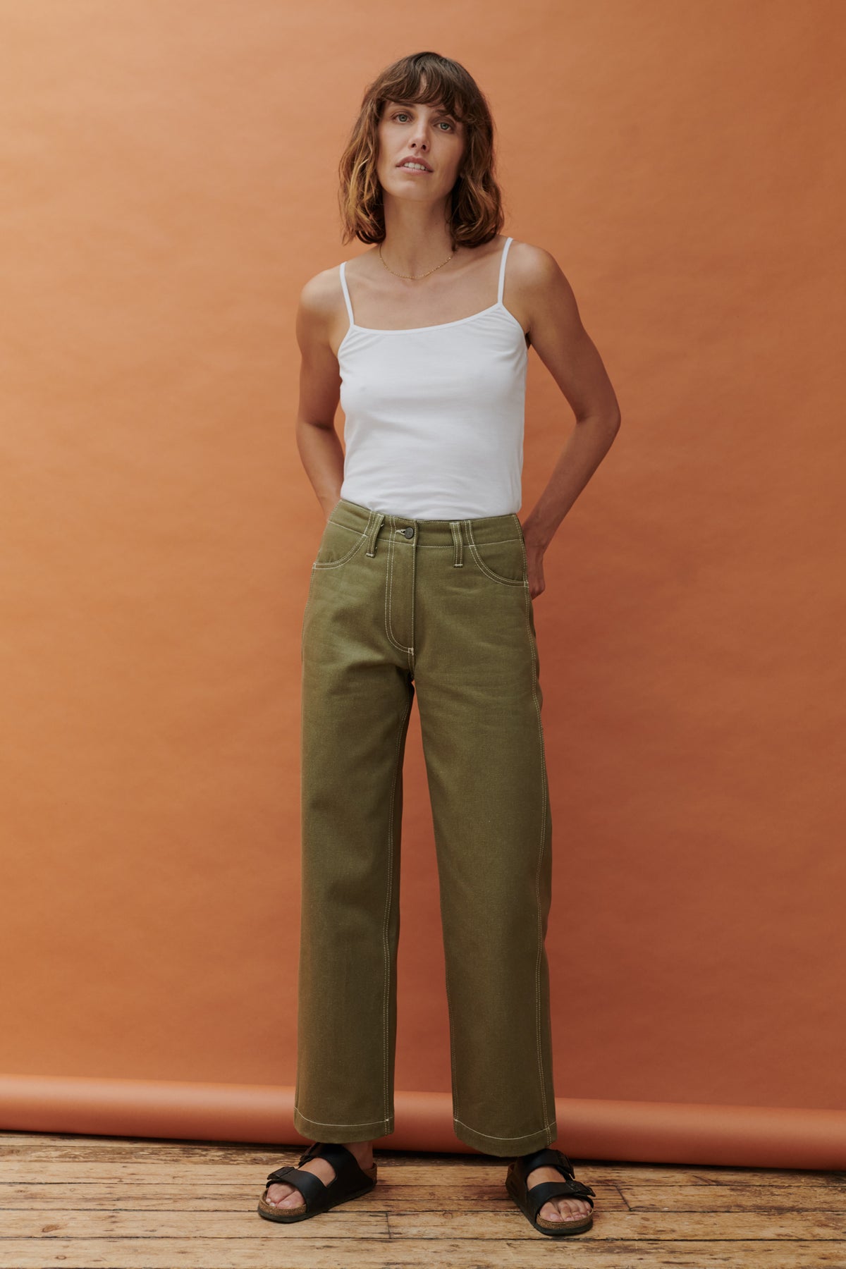 female standing in front of apricot background with hands behind her back wearing olive work jean