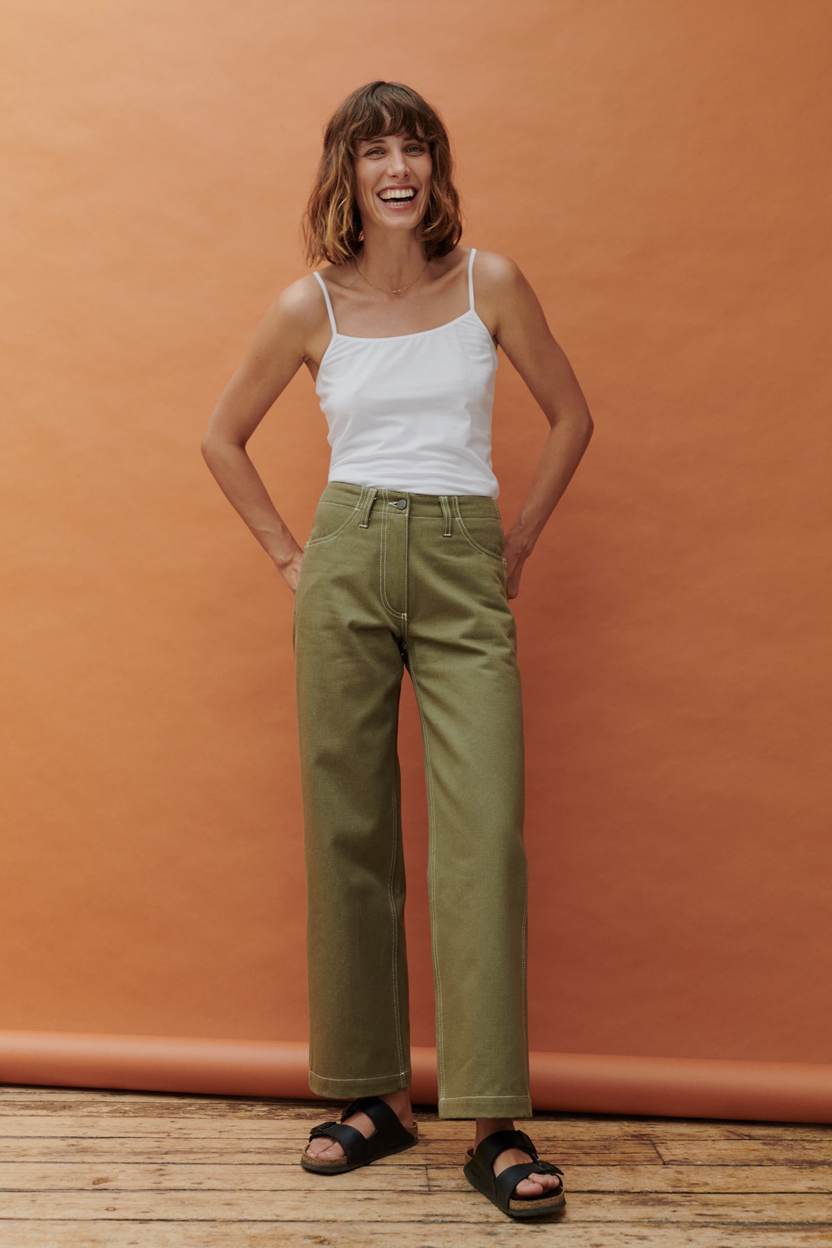 smiling female standing in front of apricot background with hands behind her back wearing olive work jean