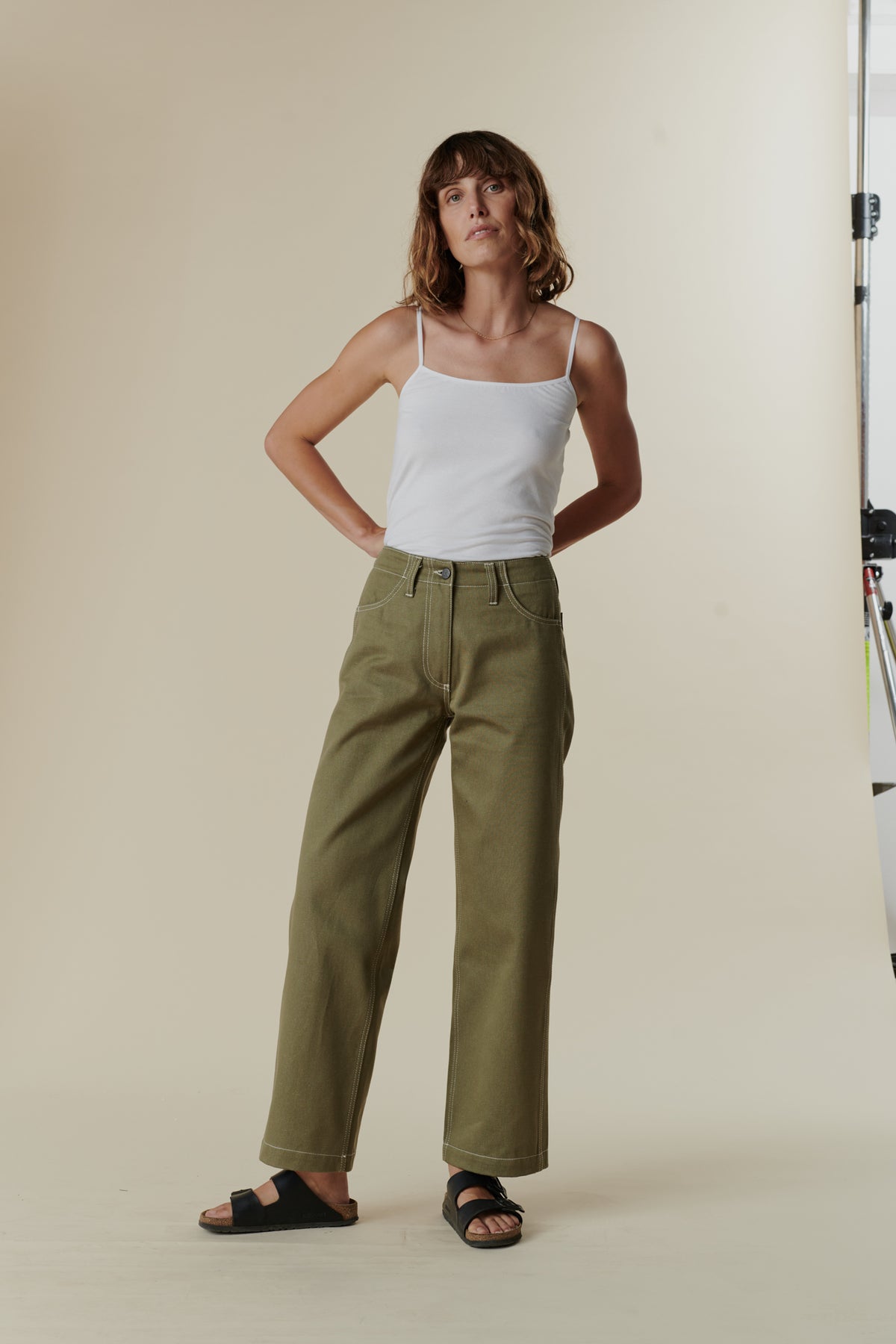 female standing in front of ecru background with hands behind her back wearing olive work jean