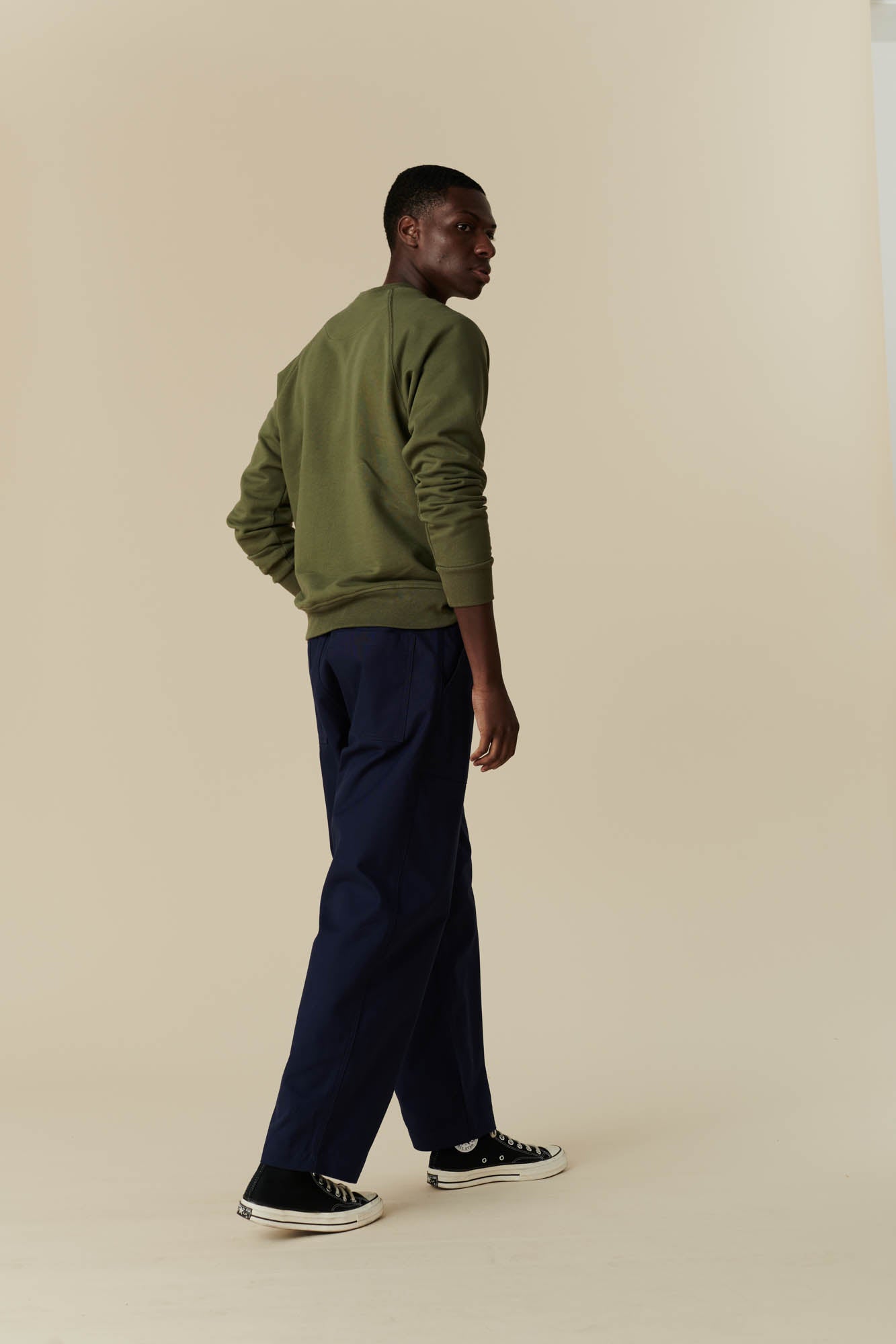 Back angle picture of a man wearing olive sweatshirt with navy tapered fit cameraman pant with black 70s Converse.