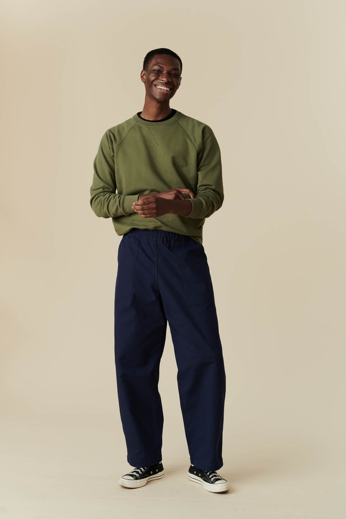 Short haired, black man wearing olive raglan sweatshirt with navy tapered fit cameraman pant with black 70s Converse.