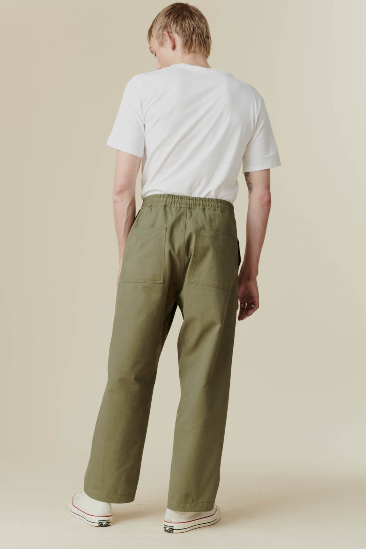 back shot of blonde male wearing olive green cameraman pant with a tucked in white t-shirt and a pair of 70s Converse trainers with a beige background