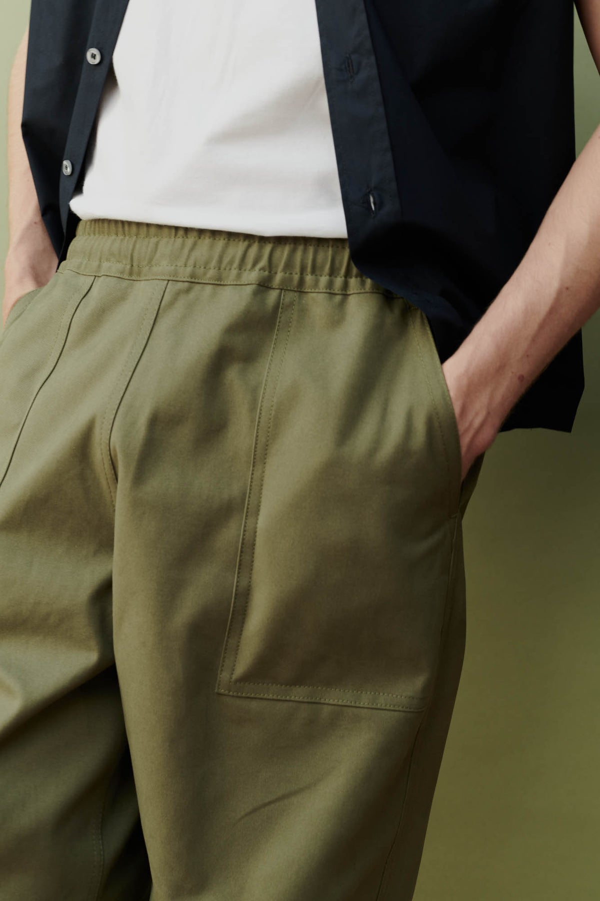 
            close up detail shot of the front left pocket of the olive green cameraman pant patch pocket. Man with hands in his pockets with an unbuttoned navy shirt.