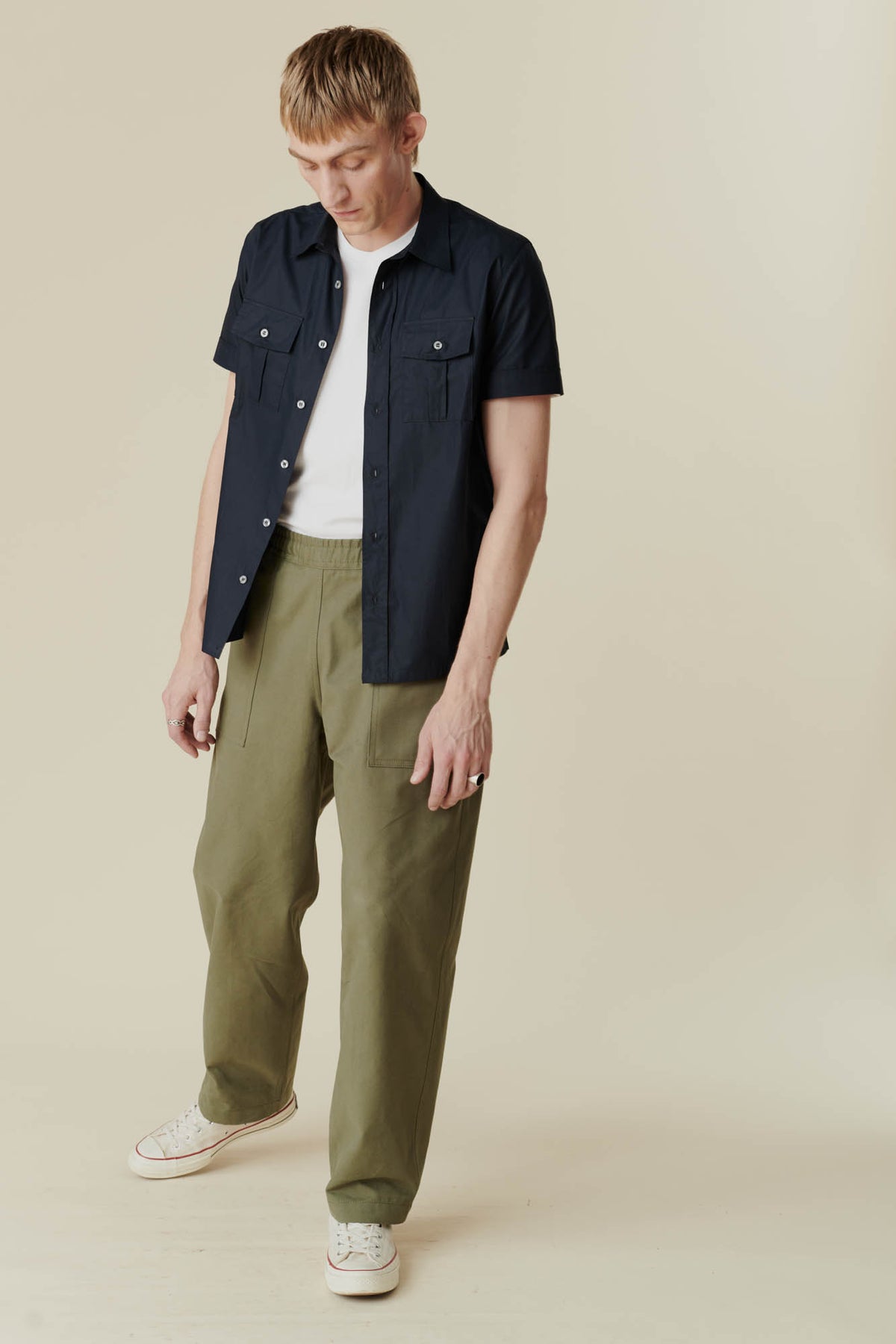 man looking down wearing olive green cameraman pant with a short sleeve navy shirt and a white t-shirt with white converse 70s trainers.