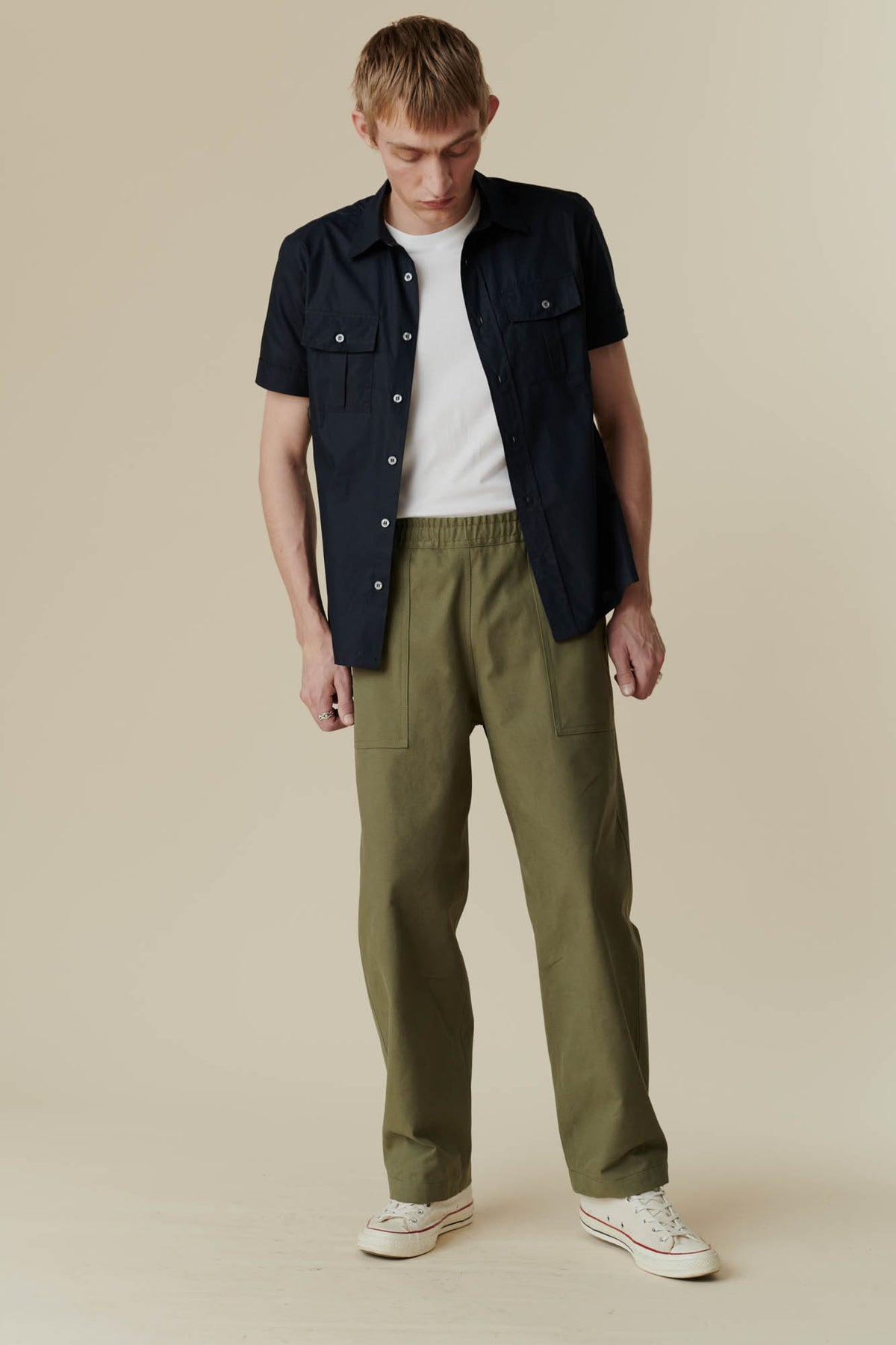 man looking down wearing olive green cameraman pant with a short sleeve navy shirt and a white t-shirt with white converse 70s trainers.