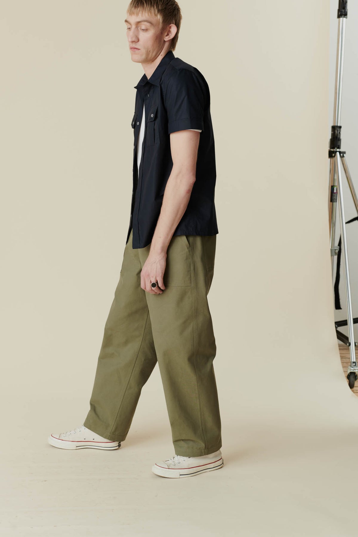 
            Side angle view of a man wearing olive green cameraman pant with a short sleeve navy shirt and a white t-shirt with white converse 70s trainers.