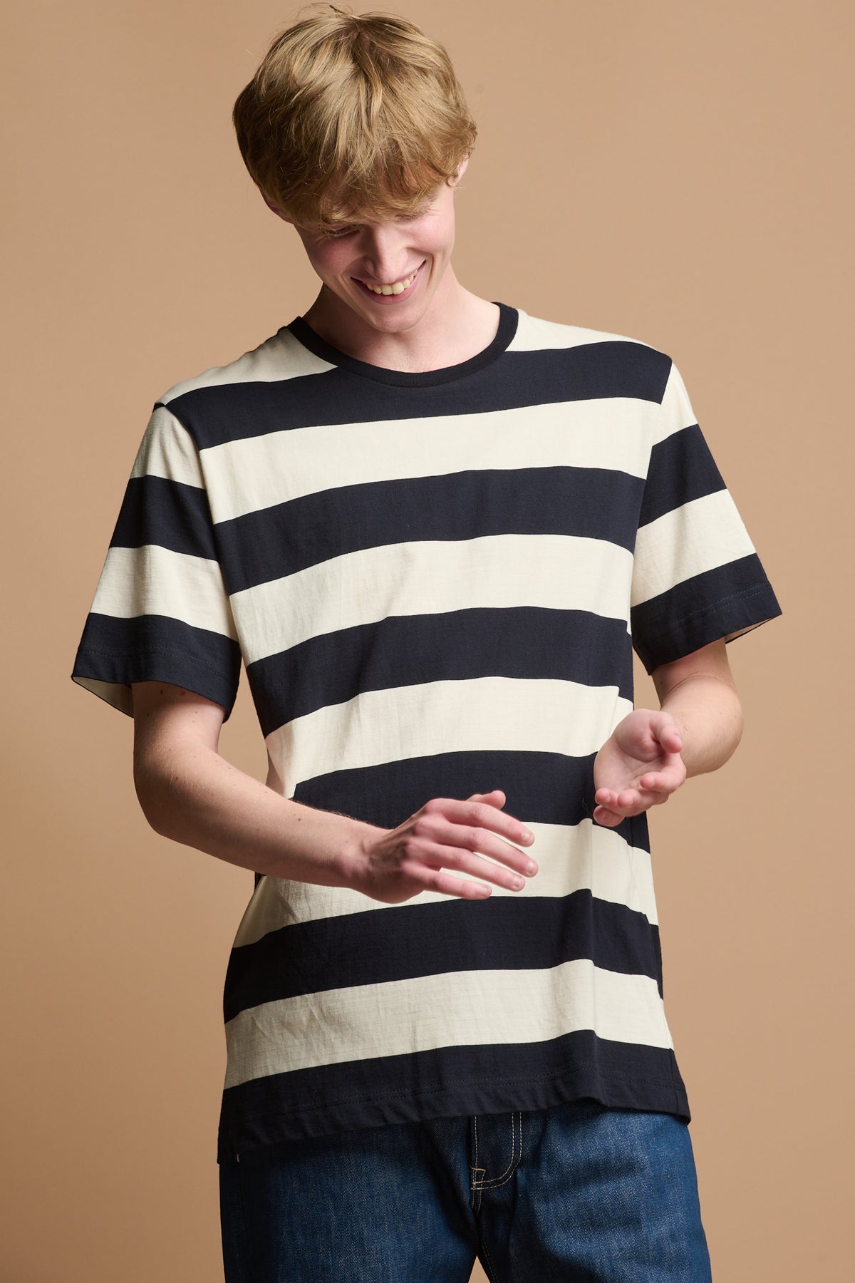 Thigh up of smiley male looking down to his hands wearing short sleeved t shirt in ecru navy wide stripe