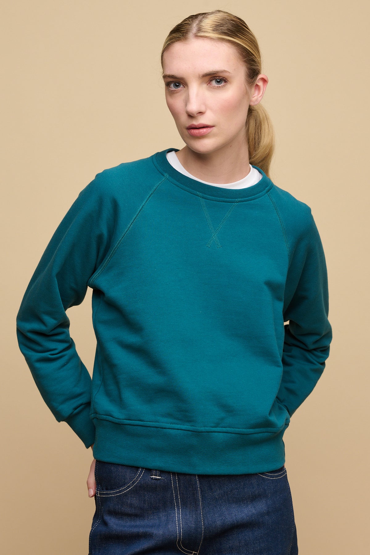 Thigh up front of female wearing Raglan sweatshirt in teal layered over crew neck t shirt in white worn with blue work jean