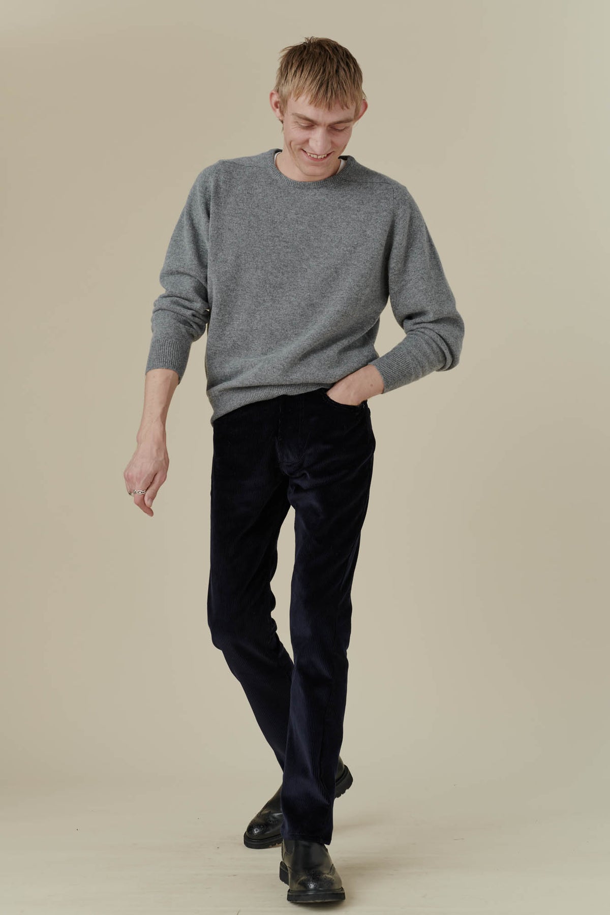 Smiley, blonde, white male wearing five pocket cord trousers in navy, with hand in front pocket, paired with grey lambswool crew neck jumper