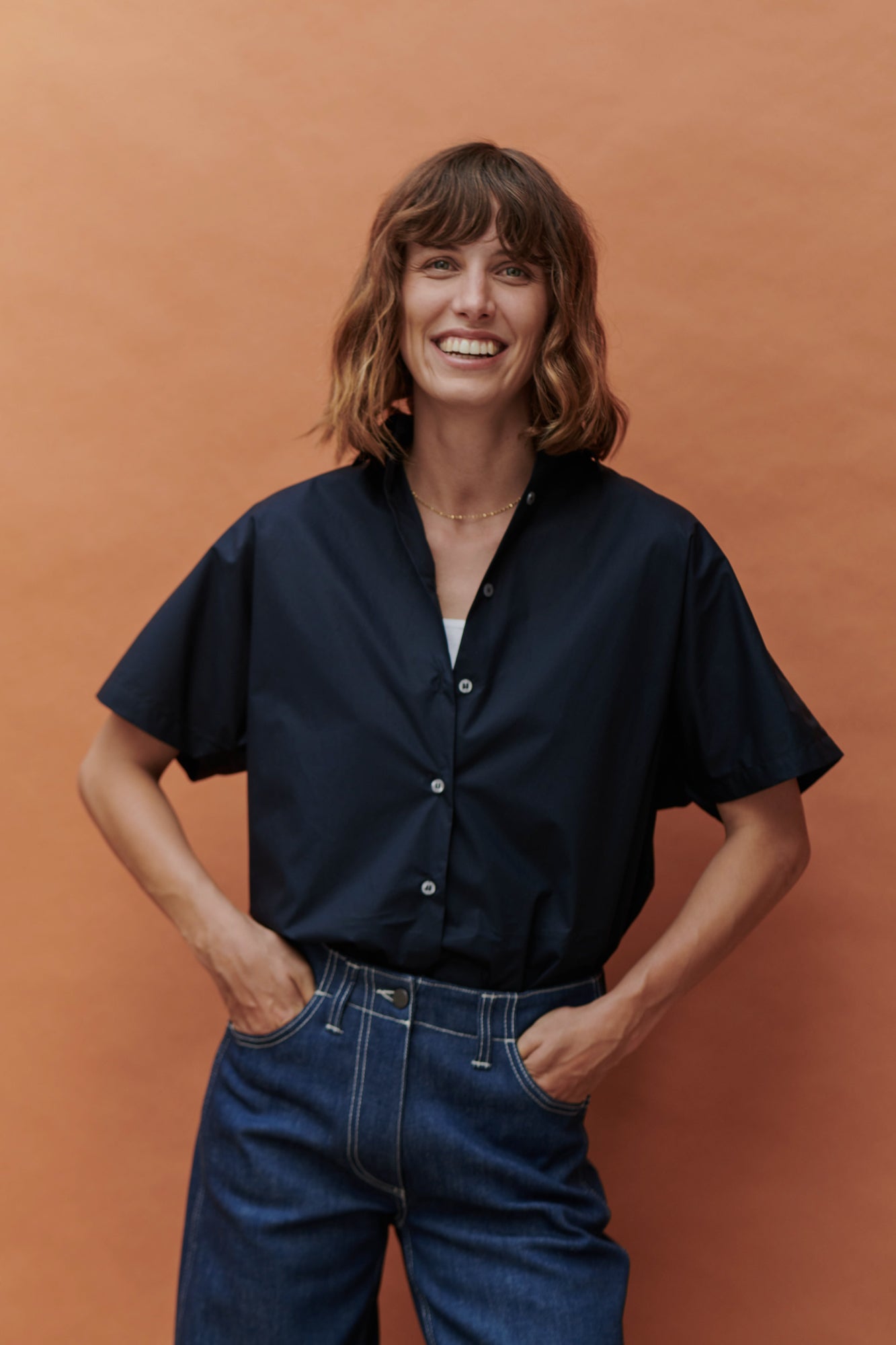 Smiling woman in front of an Apricot background smiling with her hands in her pockets of her Work Jeans and also wearing a short sleeve navy Ava shirt from Community Clothing.