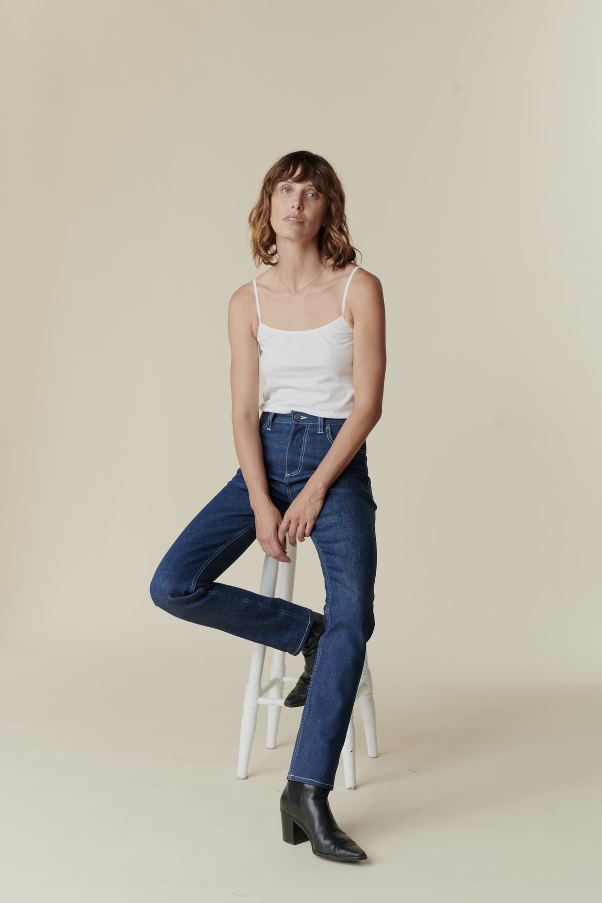 Front full body image of female wearing straight leg high rise jean in blue sat of tall stool.