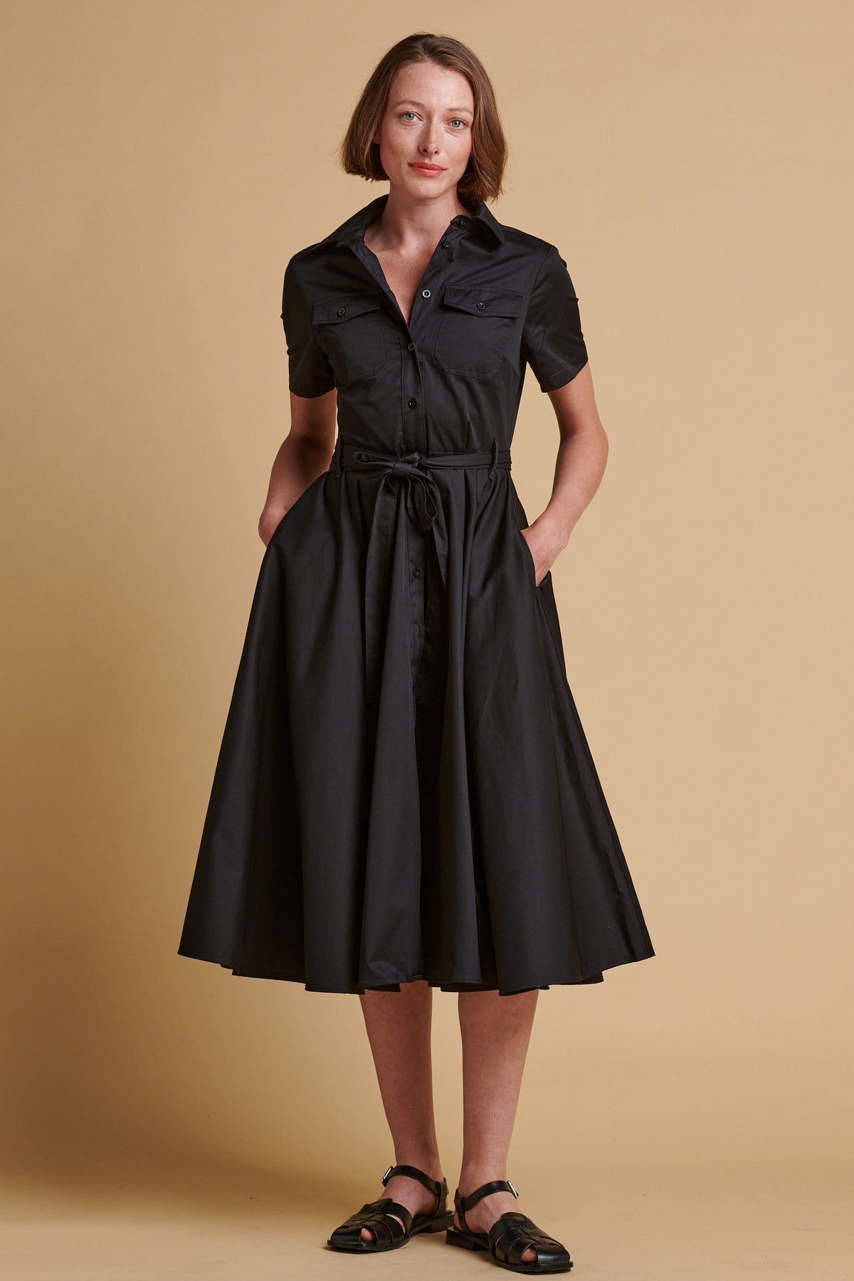 Female wearing full skirted phoebe trench dress in black with her hands in the pockets.