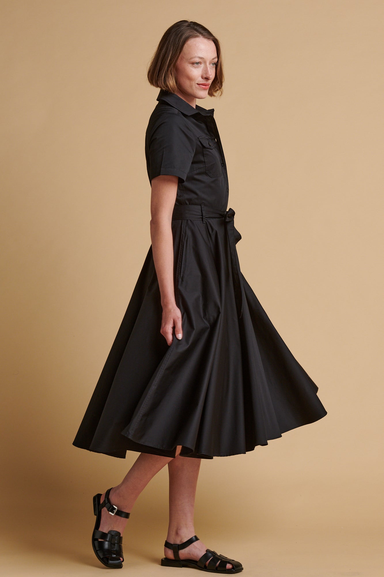 The side of female wearing full skirted phoebe trench dress in black.