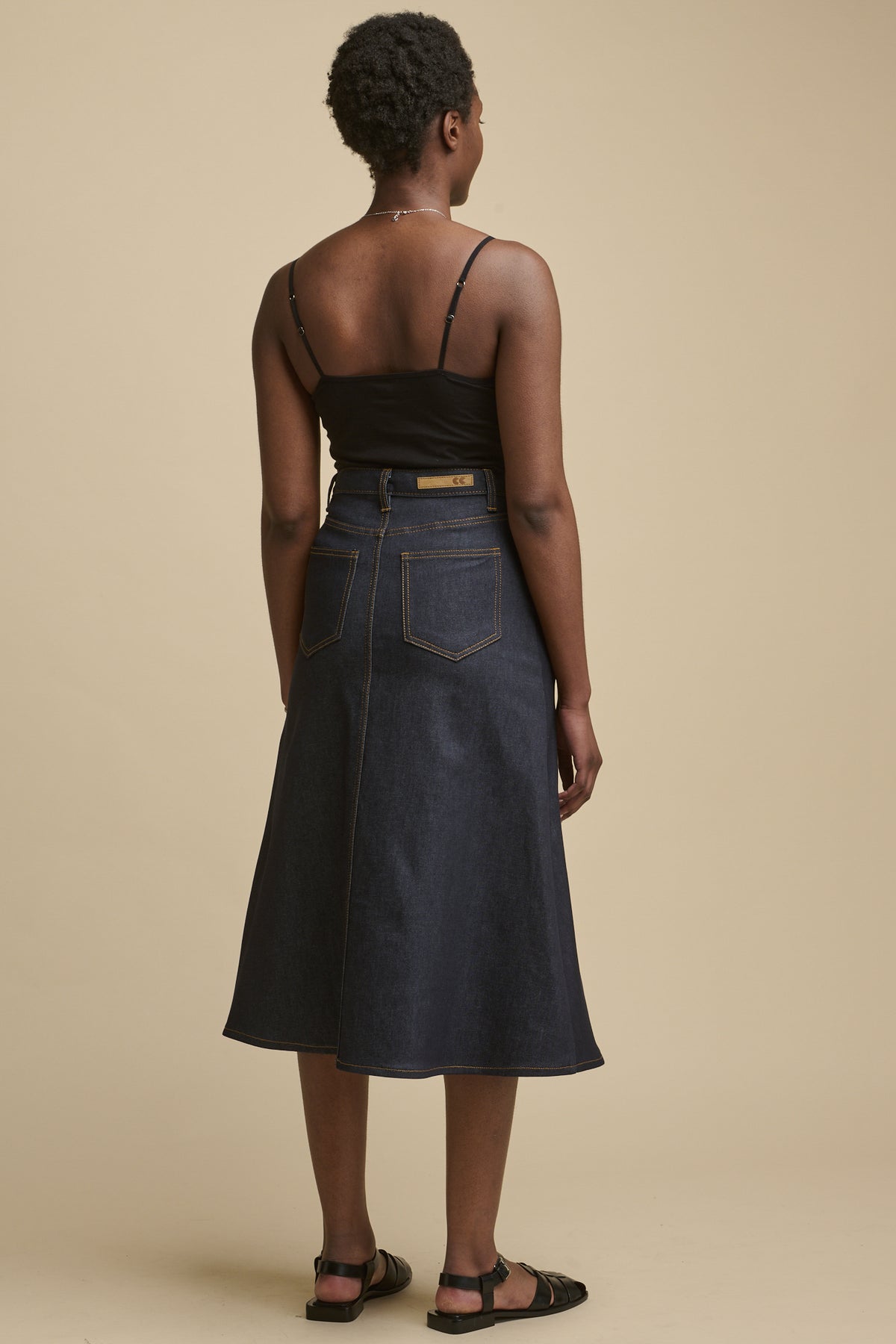 The back of female wearing Bobbie a line denim skirt in indigo paired with camisole in black and black sandals