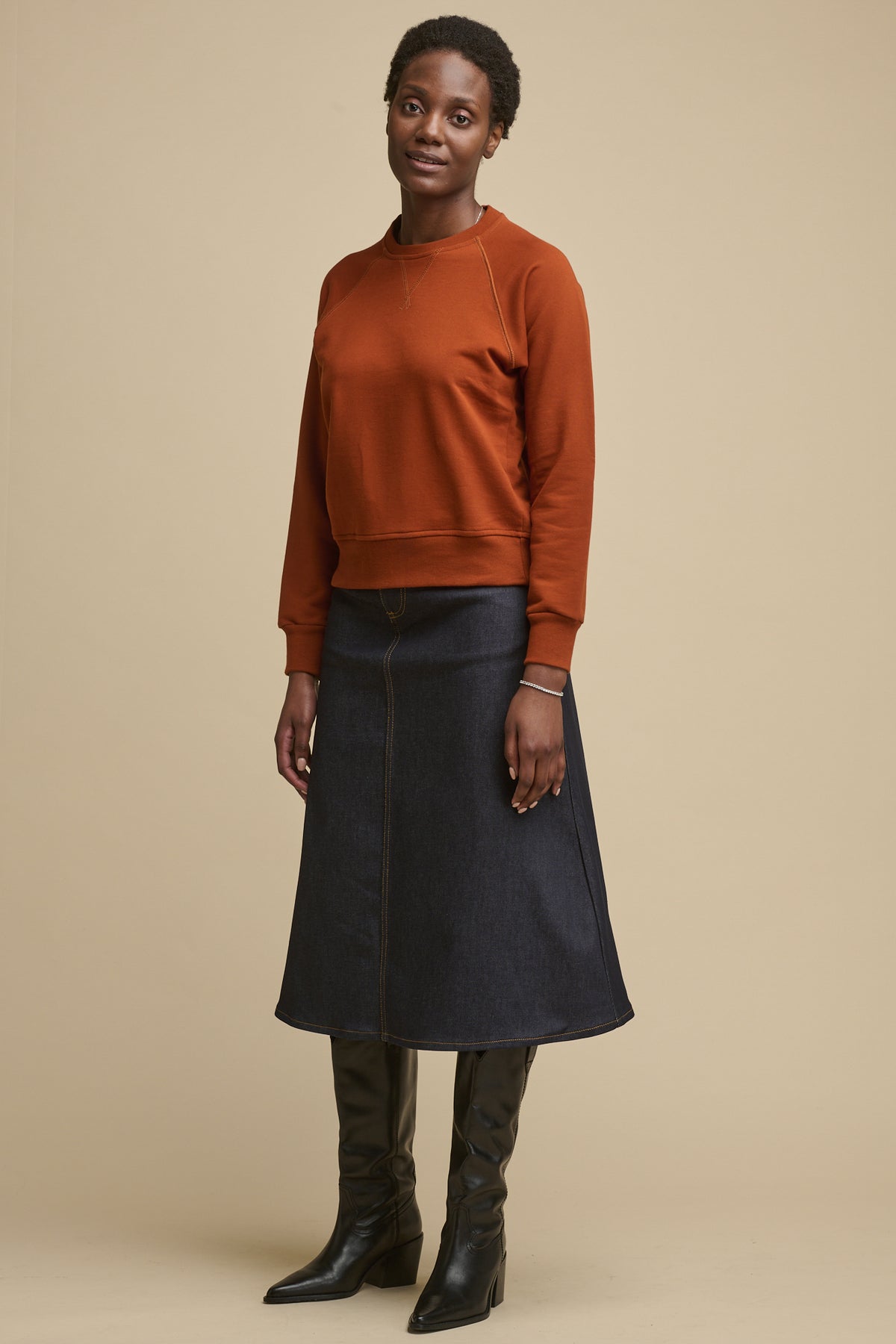 Full body image of the front of female wearing Community Clothing Bobbie a line denim skirt in indigo paired with cinnamon raglan sweatshirt