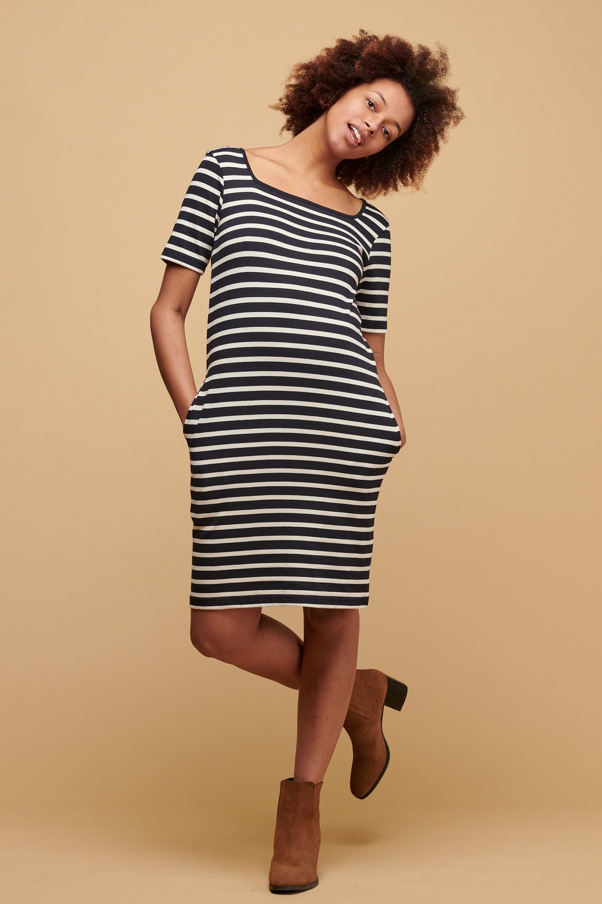 Female of colour with afro tilting her head to the side wearing navy ecru stripe Breton dress with navy binding round the square neck.