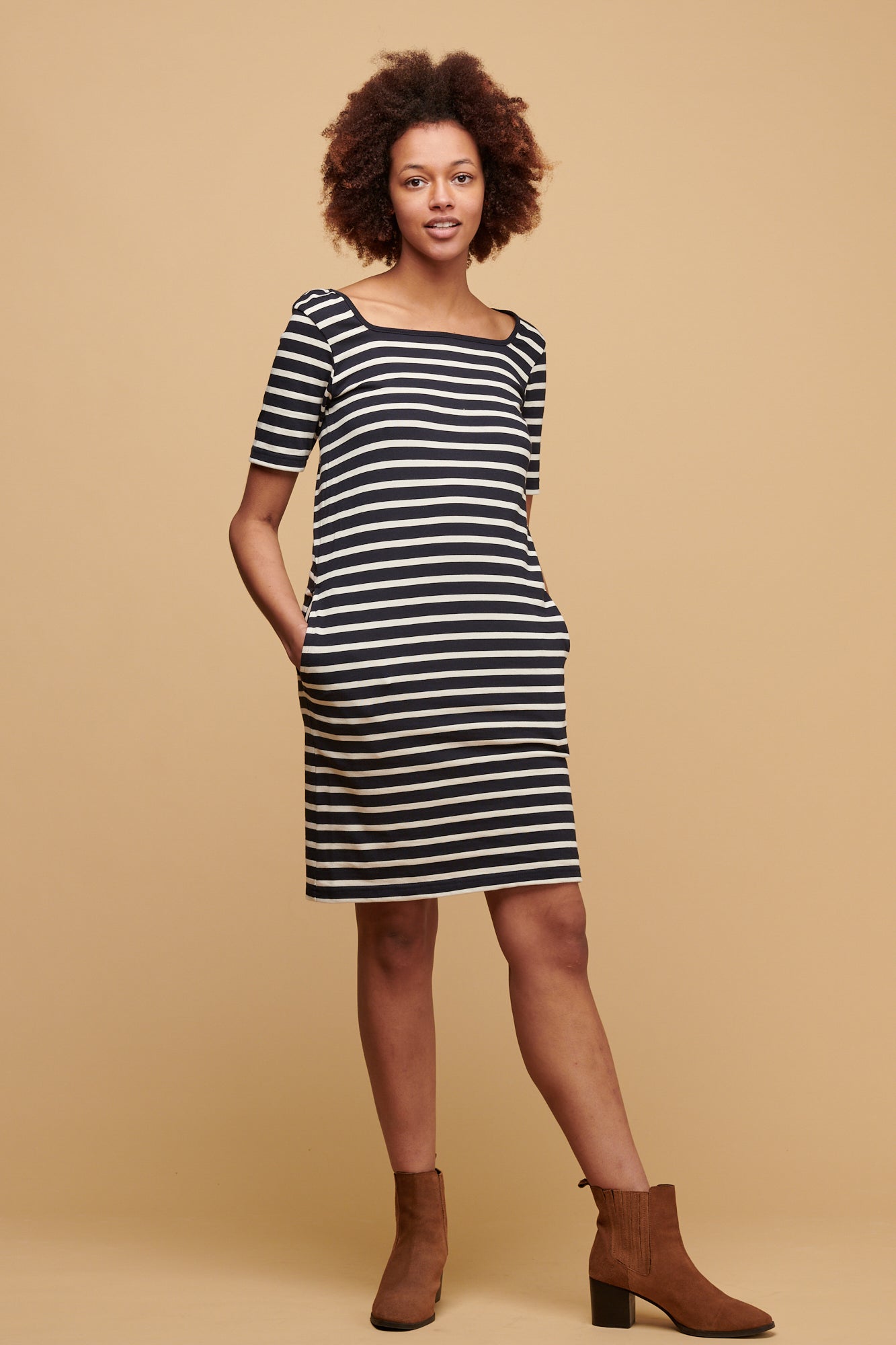 Female of colour with afro wearing navy ecru stripe Breton dress with navy binding round the square neck, with her hands in the pockets.