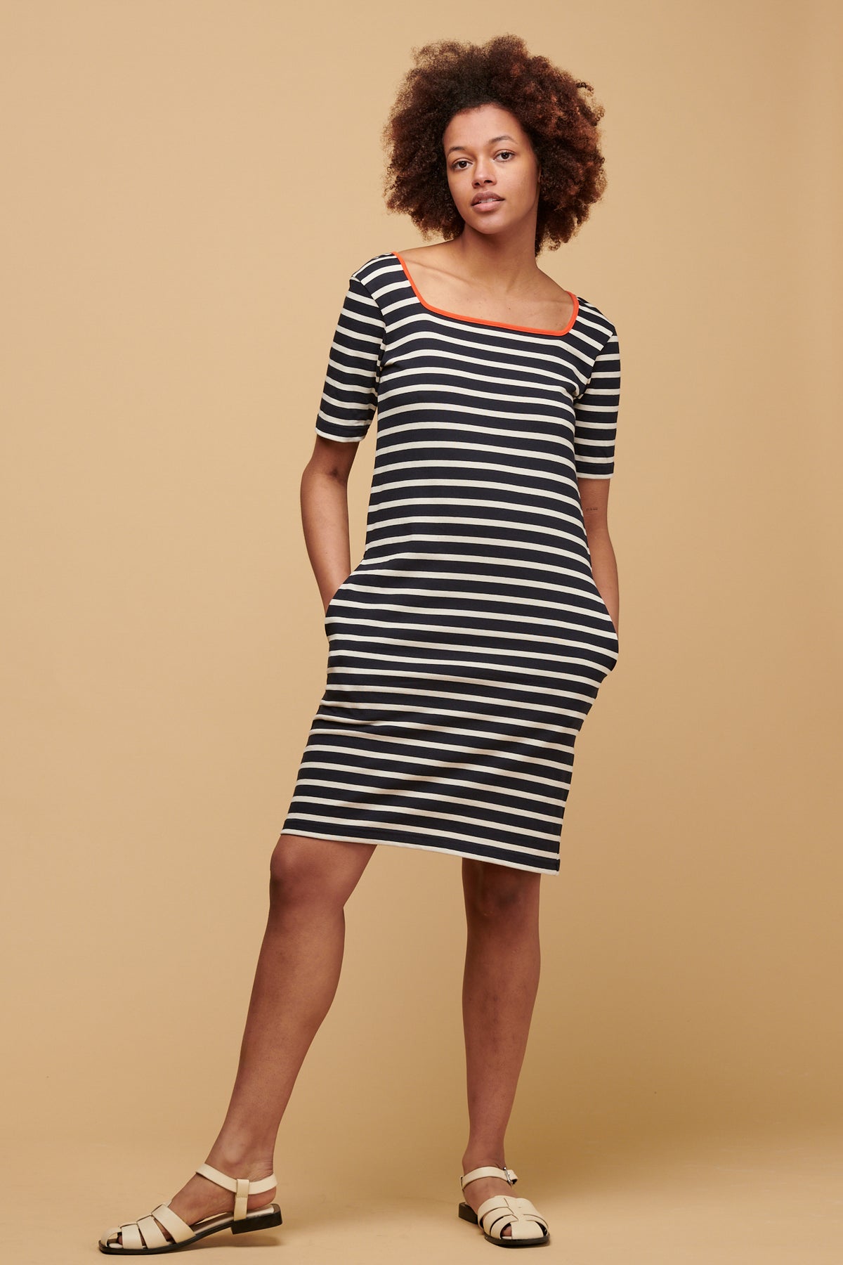 The front of female of colour wearing Breton dress in navy ecru stripe with flame red binding round the square neck, with her hands in the pockets.