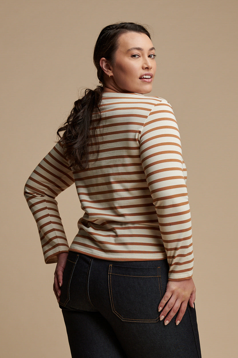 Thigh up of female from the back with brown hair tied back into a low ponytail wearing long sleeve Breton top in ecru and cinnamon stripes paired with work jean in indigo.