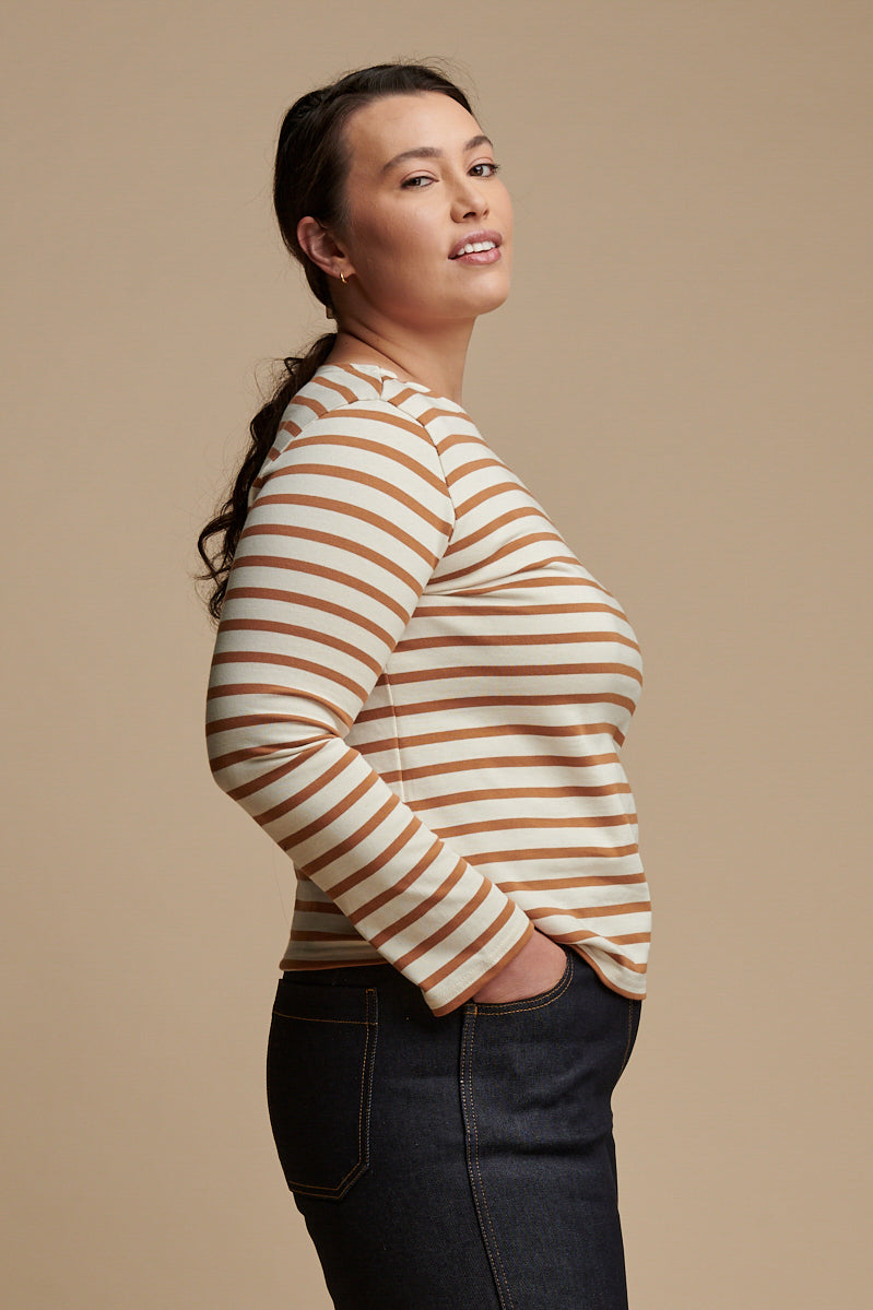 Thigh up of female from the side with brown hair tied back into a low ponytail wearing long sleeve Breton top in ecru and cinnamon stripes paired with work jean in indigo.