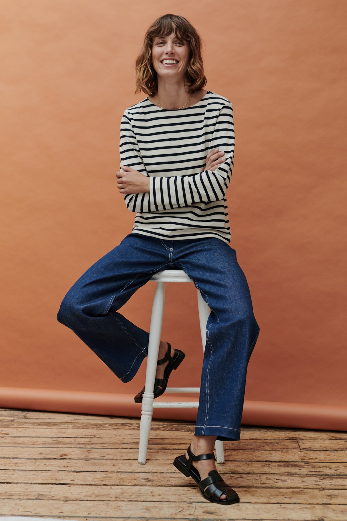 Brunette female sat on stool wearing Breton in ecru navy stripe paired with work jeans in blue