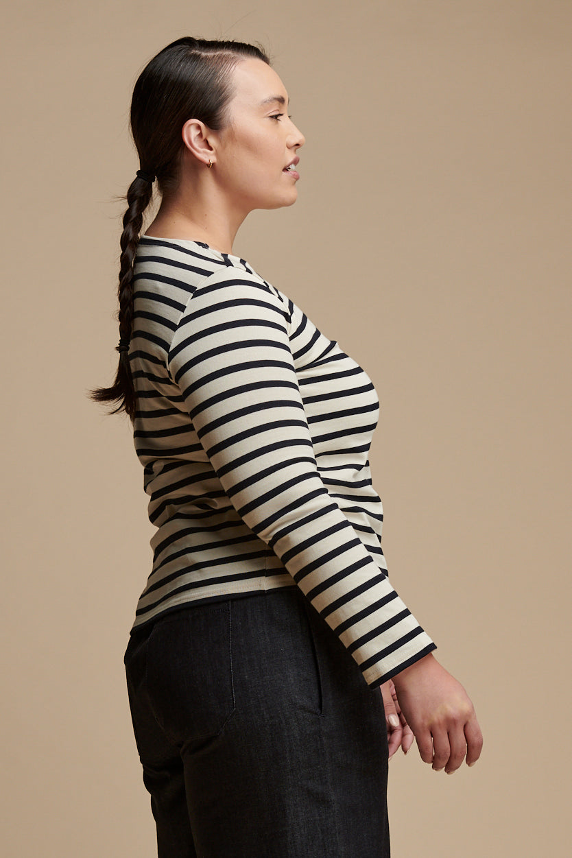 Thigh up of the side of female with hair tied back into low ponytail wearing long sleeve Breton top with boat neck in ecru navy stripes paired with indigo denim trousers.