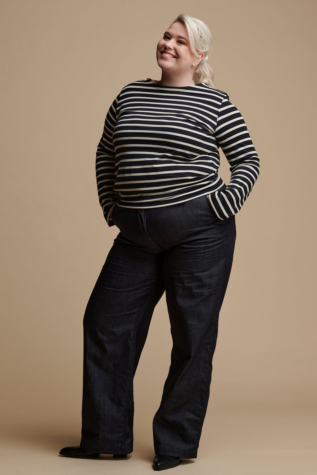 Smiley blonde female wearing navy ecru stripe Breton top paired with work trousers in indigo denim.