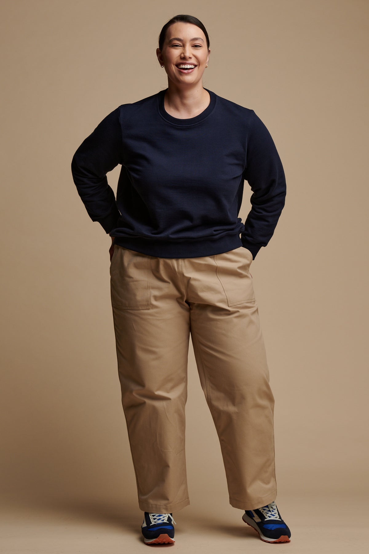 Full body image of the front of smiley female wearing camerawoman drawstring tapered cotton canvas trousers in putty paired with navy sweatshirt.
