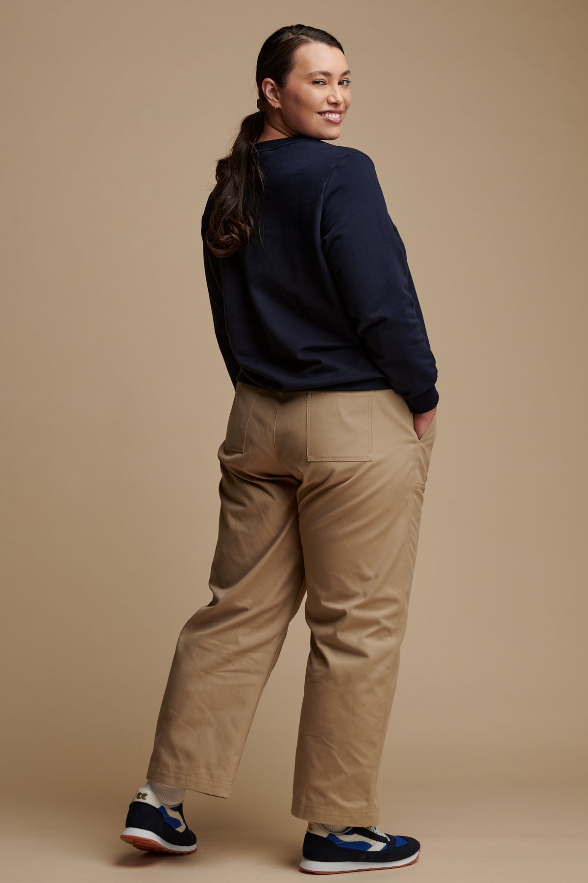 The back of smiley female wearing camerawoman drawstring tapered cotton canvas trousers in putty paired with navy sweatshirt and Community Clothing X Walsh trainers.