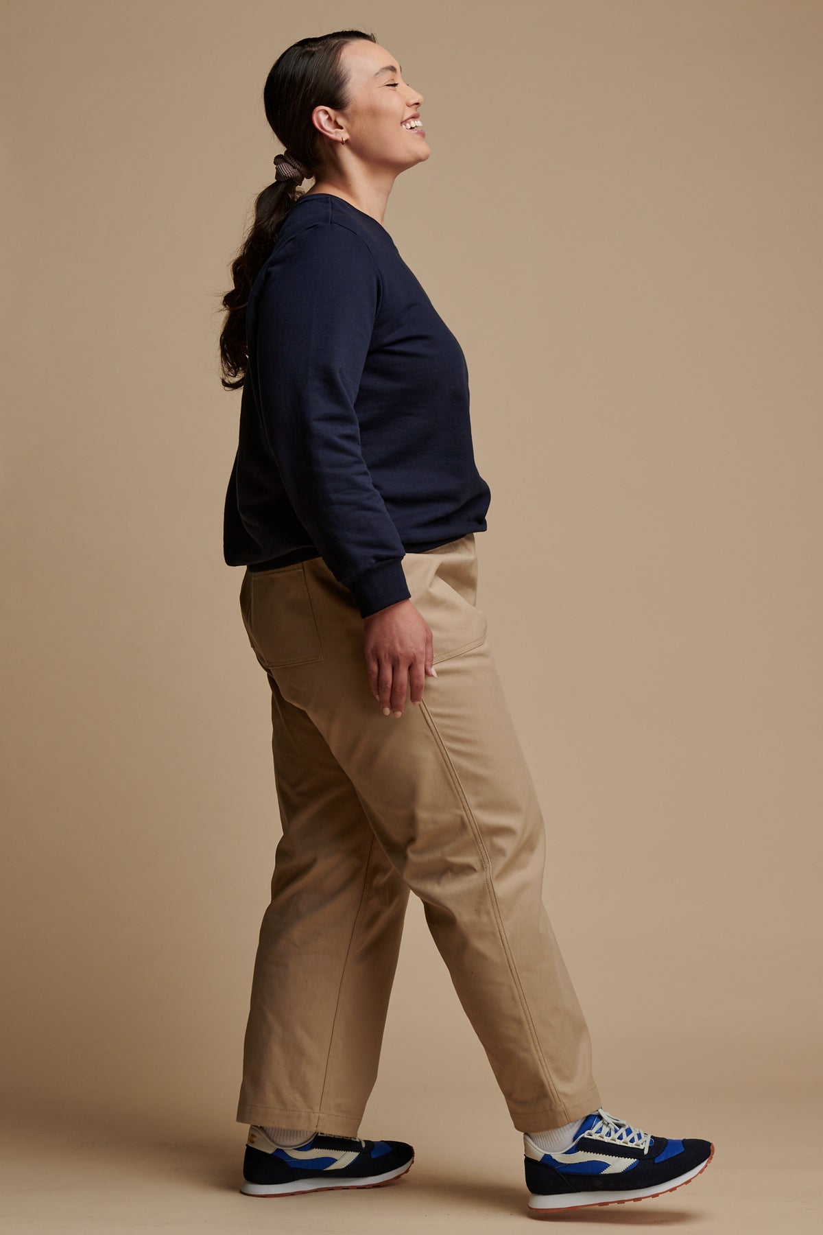 The side of smiley female wearing camerawoman drawstring tapered cotton canvas trousers in putty paired with navy sweatshirt and Community Clothing X Walsh trainers.