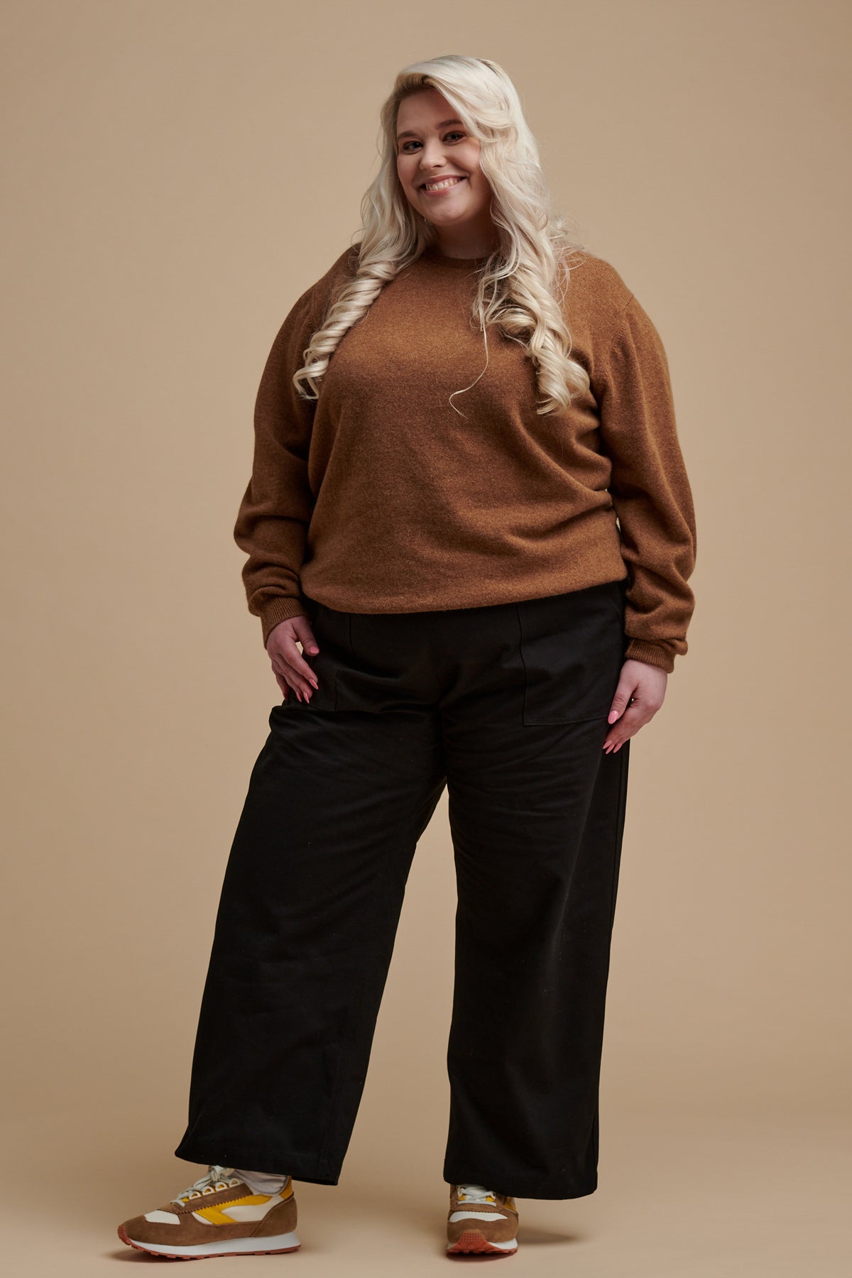 Full body of smiley female with long blonde hair wearing camerawoman drawstring tapered cotton canvas trousers in black paired with lambswool crew neck jumper in dark camel.