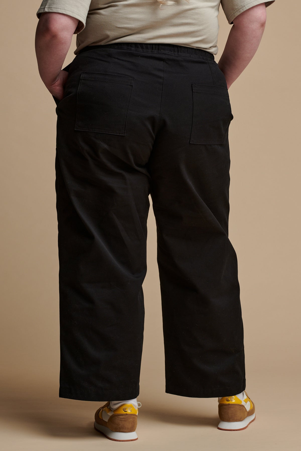 Shoulder down of the back of female wearing camerawoman drawstring tapered cotton canvas trousers in black paired with Walsh x Community Clothing trainers.